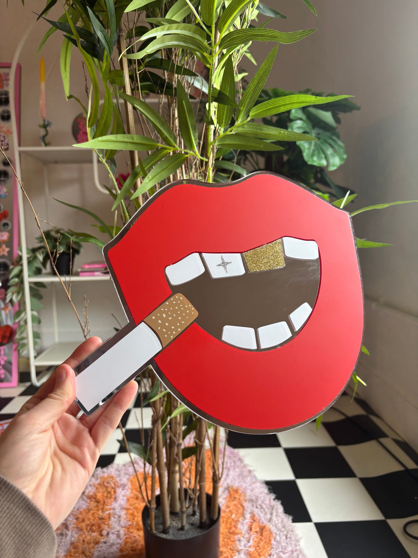 A hand holding a red and white sign with a graphic of a mouth and a cigarette, with a plant in the background.