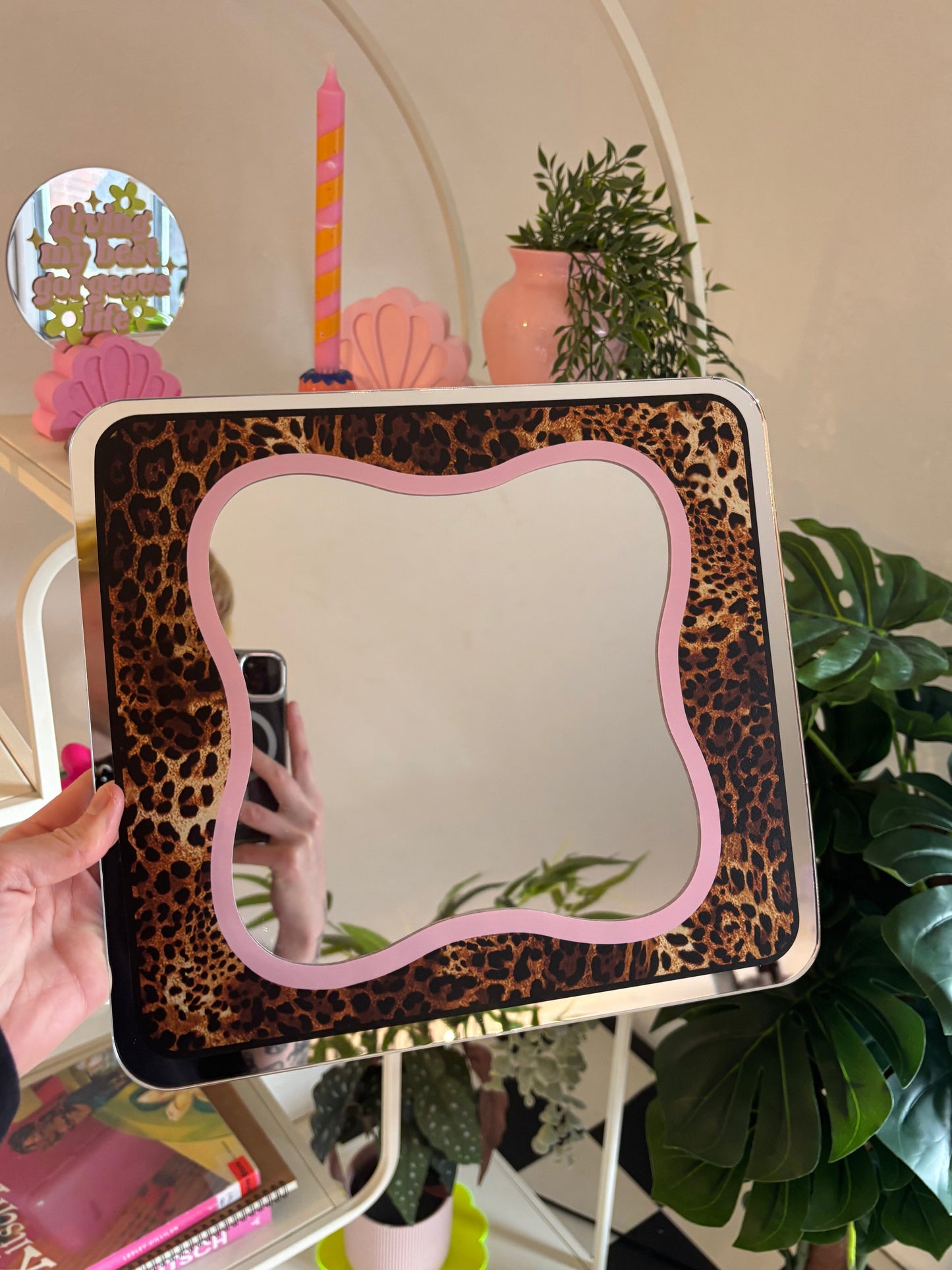 A hand is holding a leopard print-patterned mirror with a pink frame, which is placed on a white shelf among various decorative items such as a candle, a vase, and potted plants.