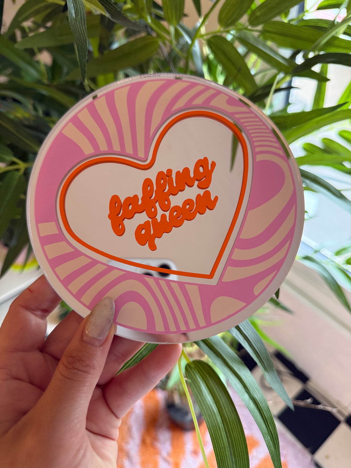 A hand holding a heart-shaped mirror with the text "I love you" written on it, against a backdrop of green leaves and a patterned wall.