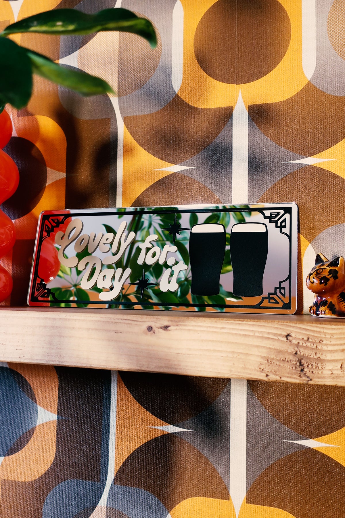 A wooden shelf displaying a sign with the text "Lovely day for beer" and two beer glasses, along with a small figurine of a cat.