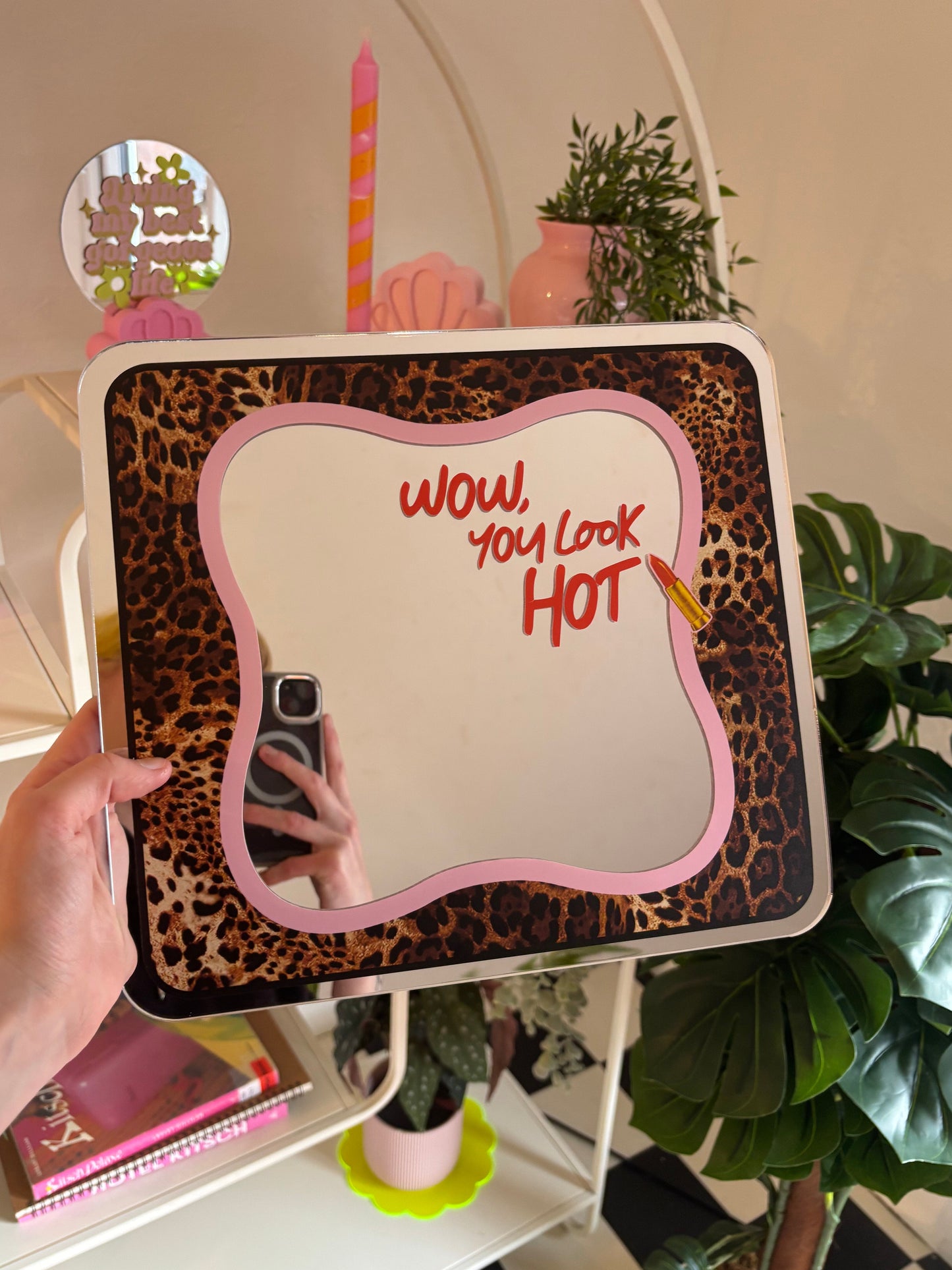 A hand is holding a mirror with a leopard print frame and the text "WOW, YOU LOOK HOT" written on it. The mirror is reflecting the hand holding it, and there are various decorative items, such as a candle, a vase, and potted plants, in the background.