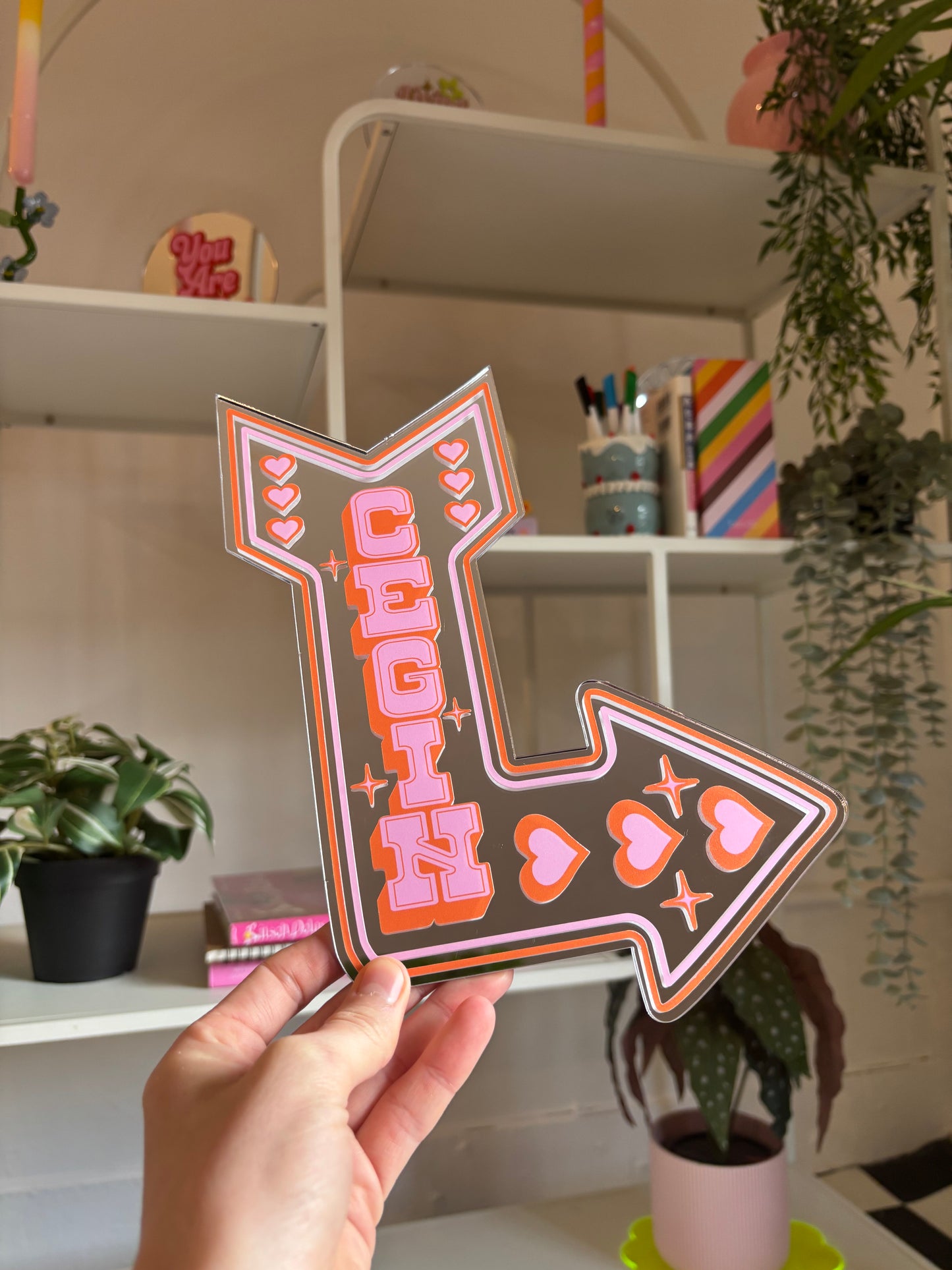 A hand holding a large, colorful sign with the name "CEGH" on it, surrounded by various decorative items such as plants and shelves.
