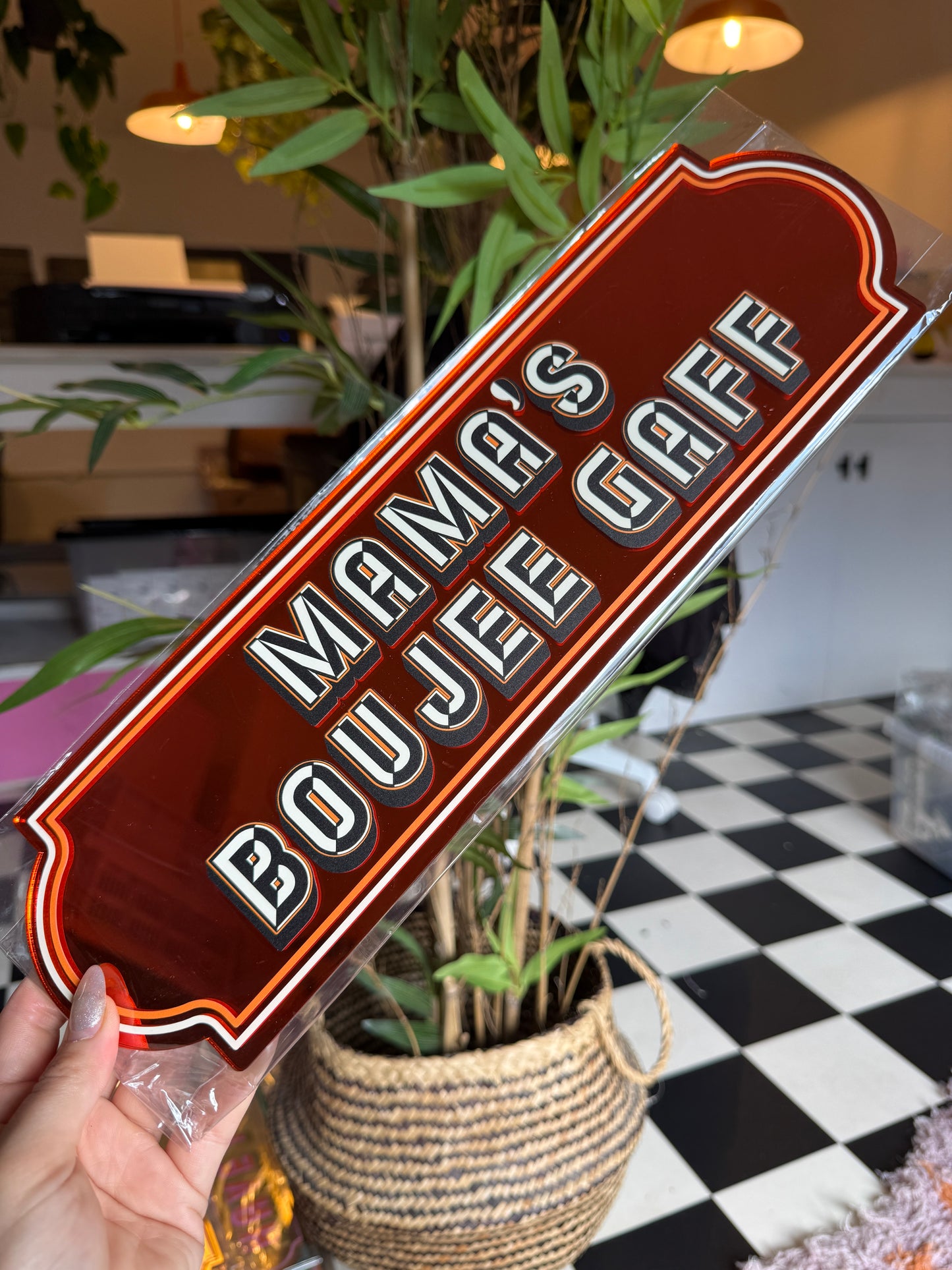 A person is holding a red sign with the text "BOUQUE S" on it, which is placed on a table with a plant and a basket.
