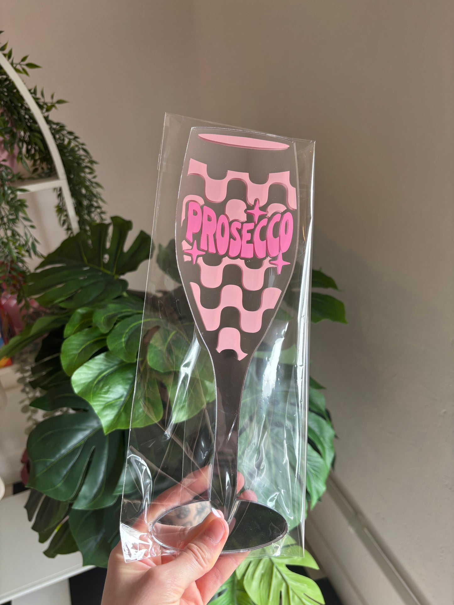 A hand is holding a clear plastic glass with a pink and black design featuring the word "PROSECCO" on it.
