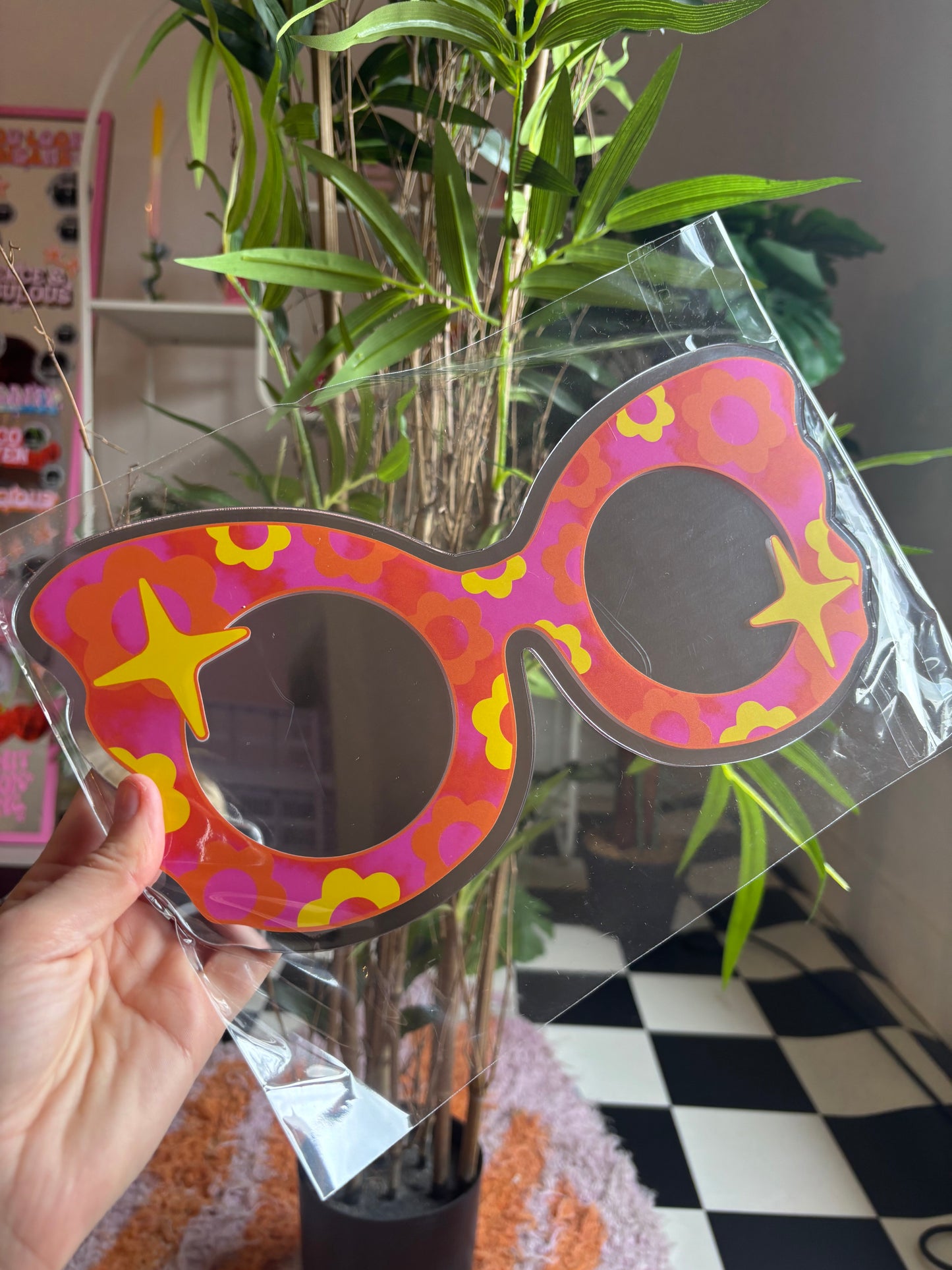 A hand is holding a colorful, patterned pair of sunglasses in front of a potted plant.