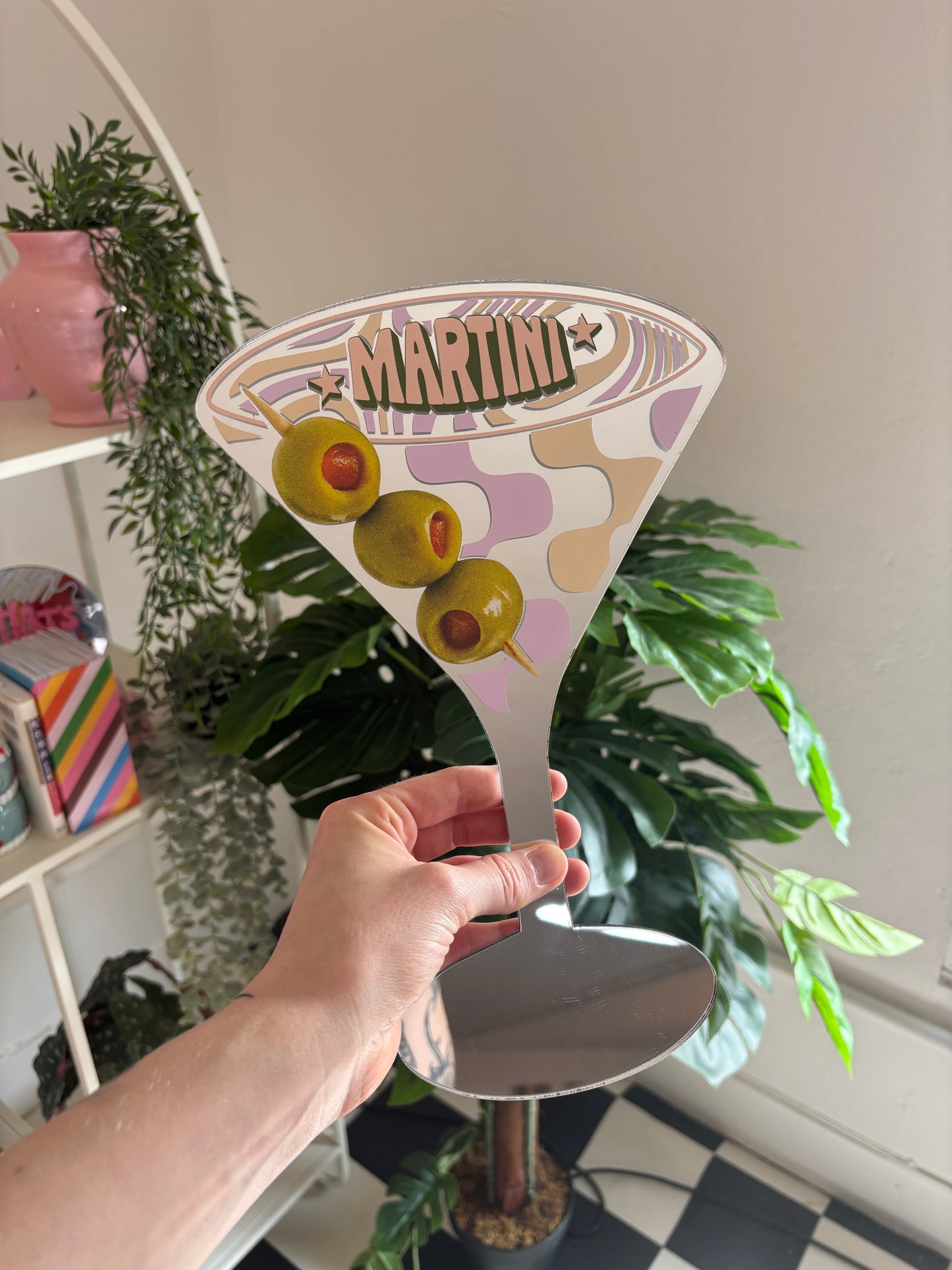 A hand is holding a decorative martini glass with a design featuring olives and a star, surrounded by potted plants and a shelf with various items.