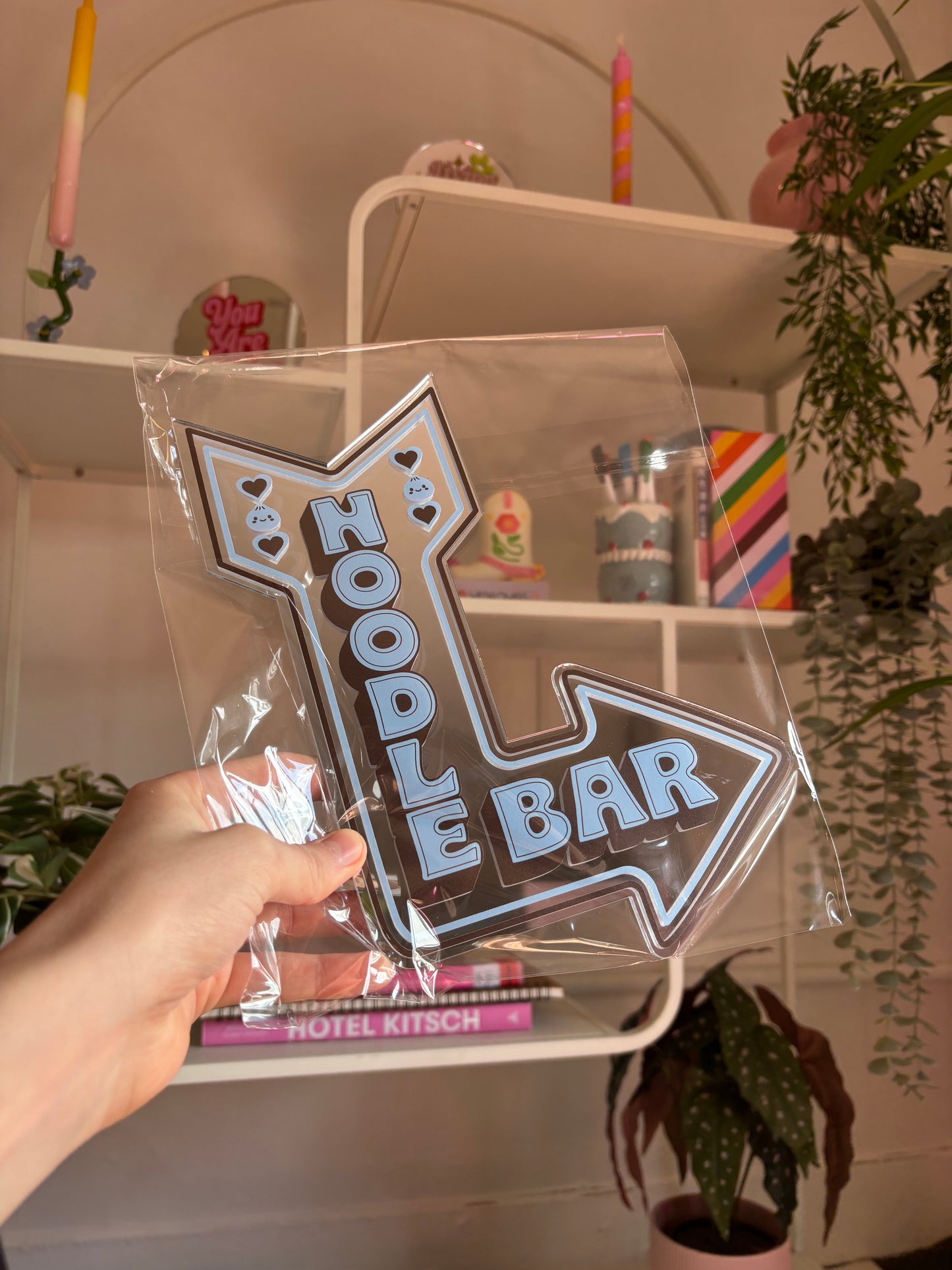 A hand is holding a clear plastic sign with the text "Noodle Bar" written on it, which is placed on a shelf among various decorative items.