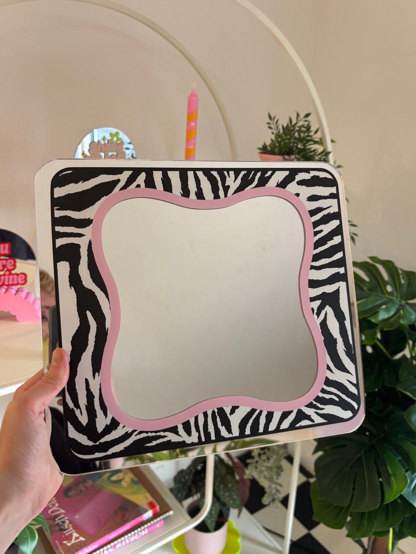 A hand is holding a square mirror with a zebra-striped frame, which is placed on a white shelf in a room with various decorative items.