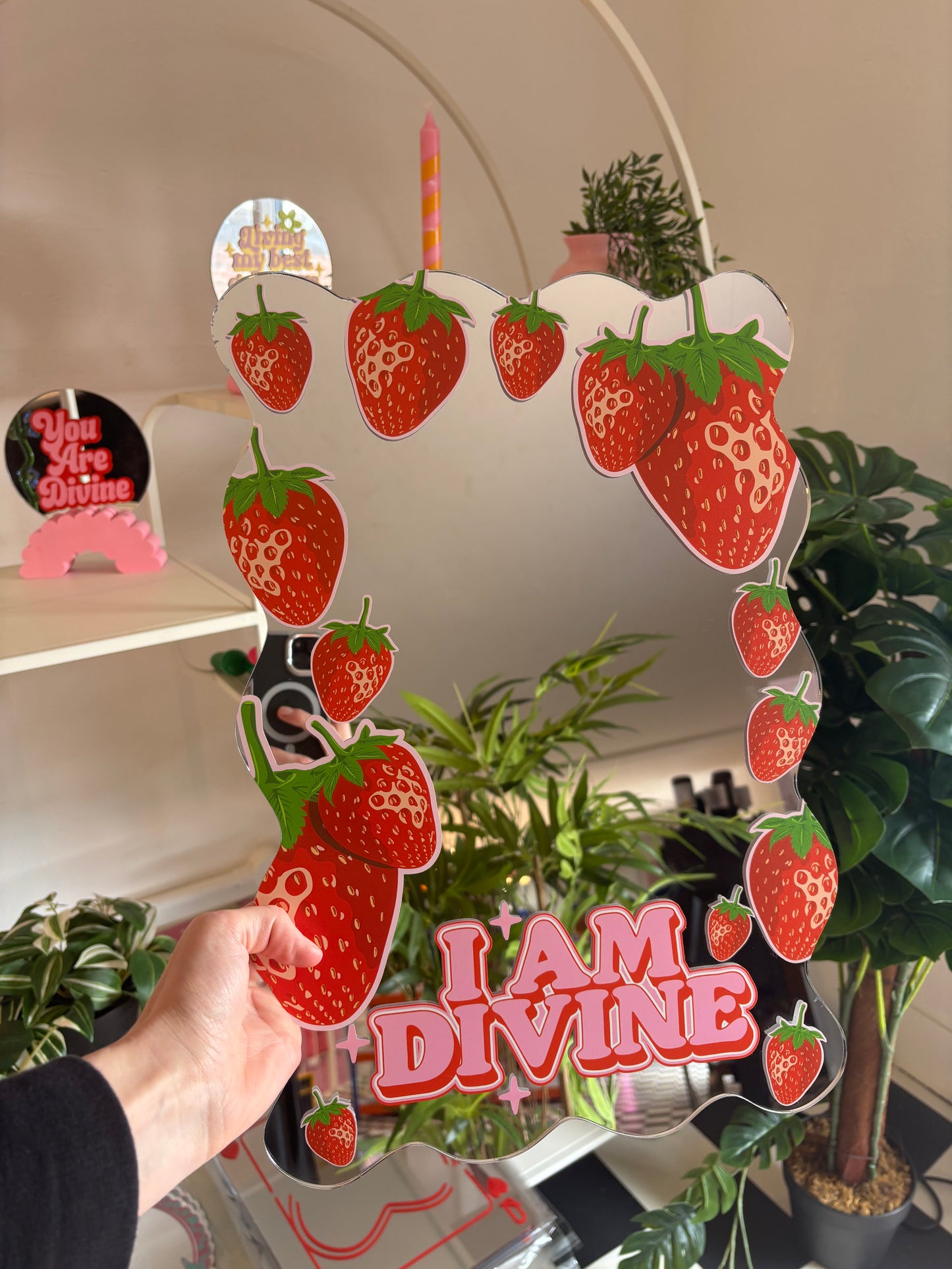 A person is holding a mirror with a frame featuring a strawberry design and the text "I am Divine".