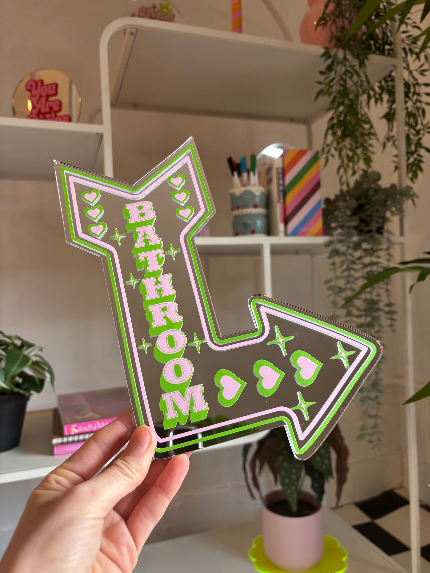 A hand is holding a large, colorful sign that says "BATHROOM" in a green and pink design, with stars and hearts around it.