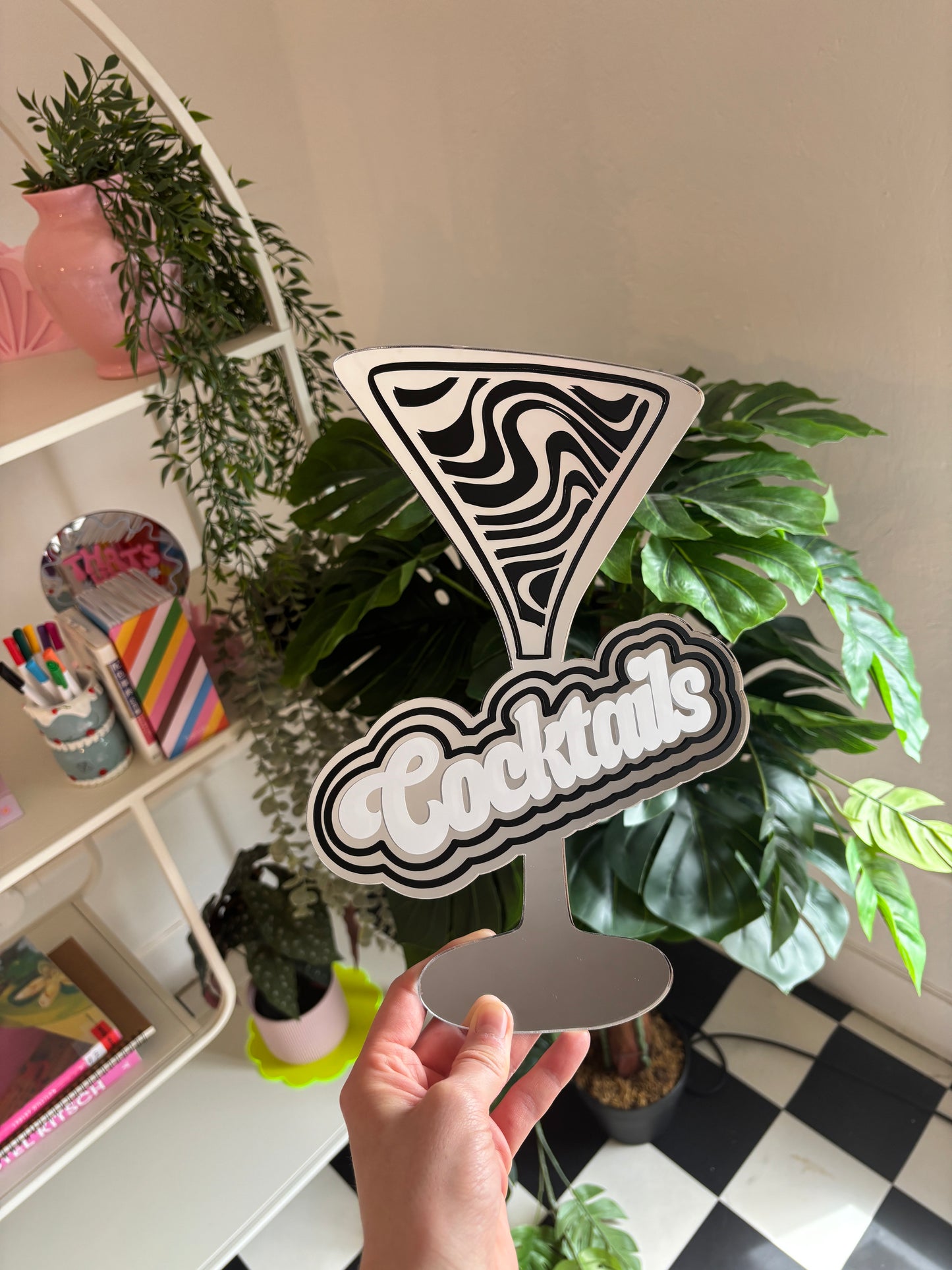 A hand is holding a cocktail glass with the word "Cocktails" written on it, surrounded by various plants and decorative items.