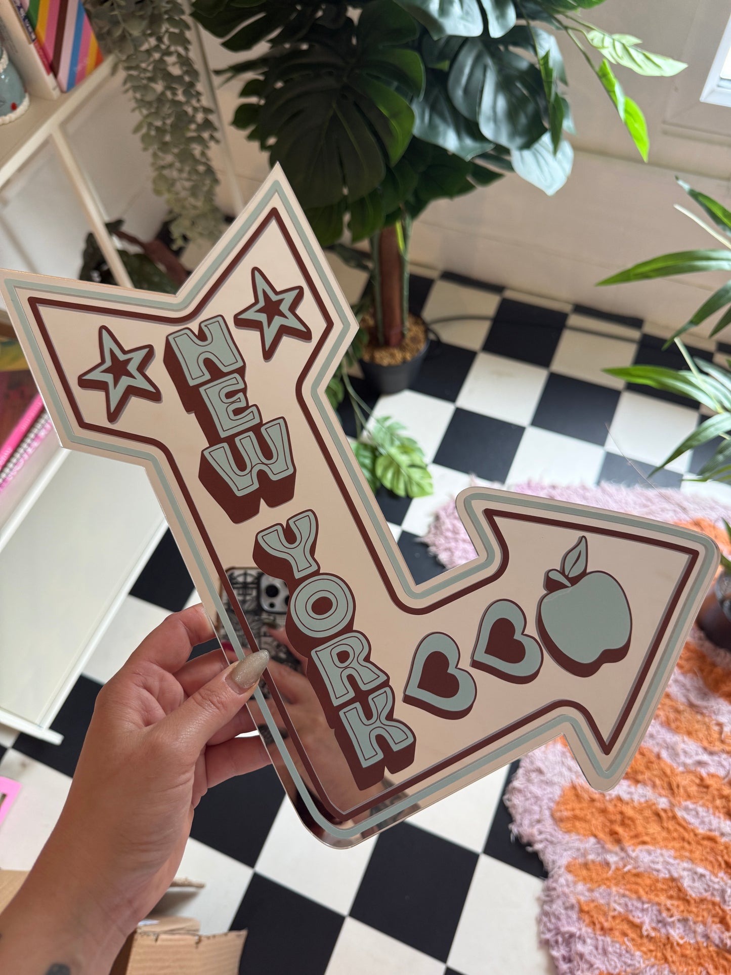 A hand is holding a sign with a stylized arrow design and various decorative elements, such as stars and hearts, against a backdrop of plants and a checkered wall.