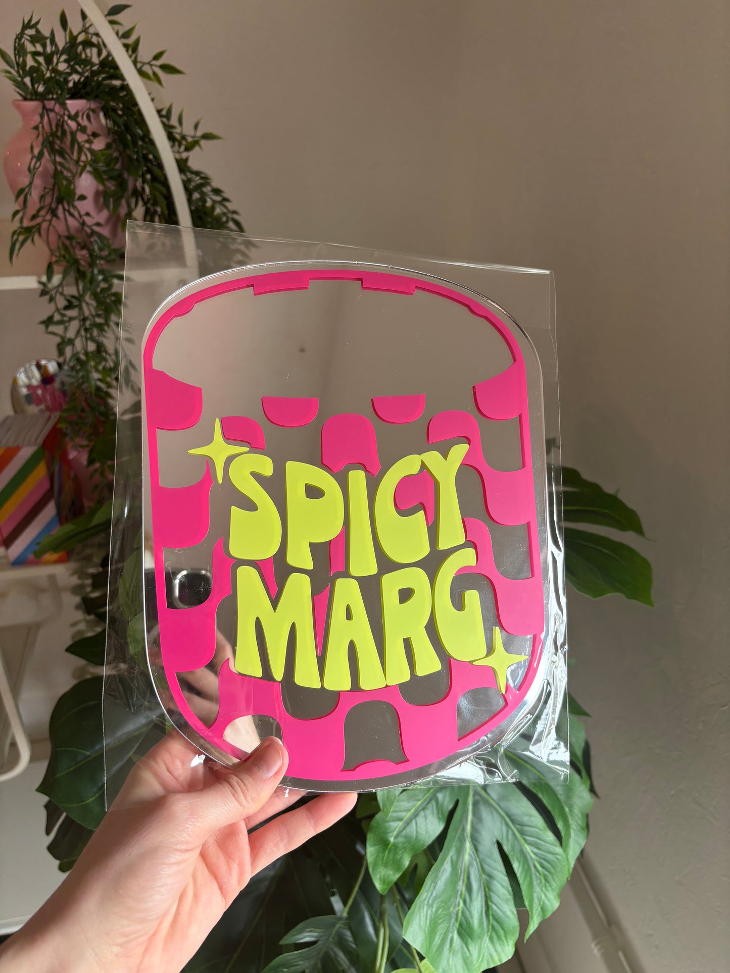 A hand is holding a pink and yellow sign with the text "SPICY MARG" against a backdrop of green plants.