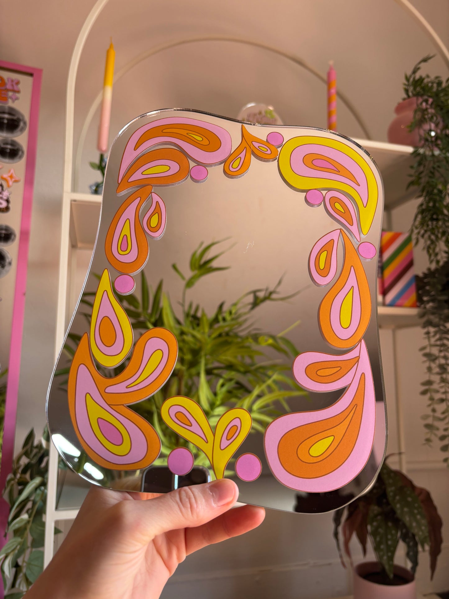 A hand is holding up a mirror with a colorful, abstract design featuring pink, orange, and yellow shapes.