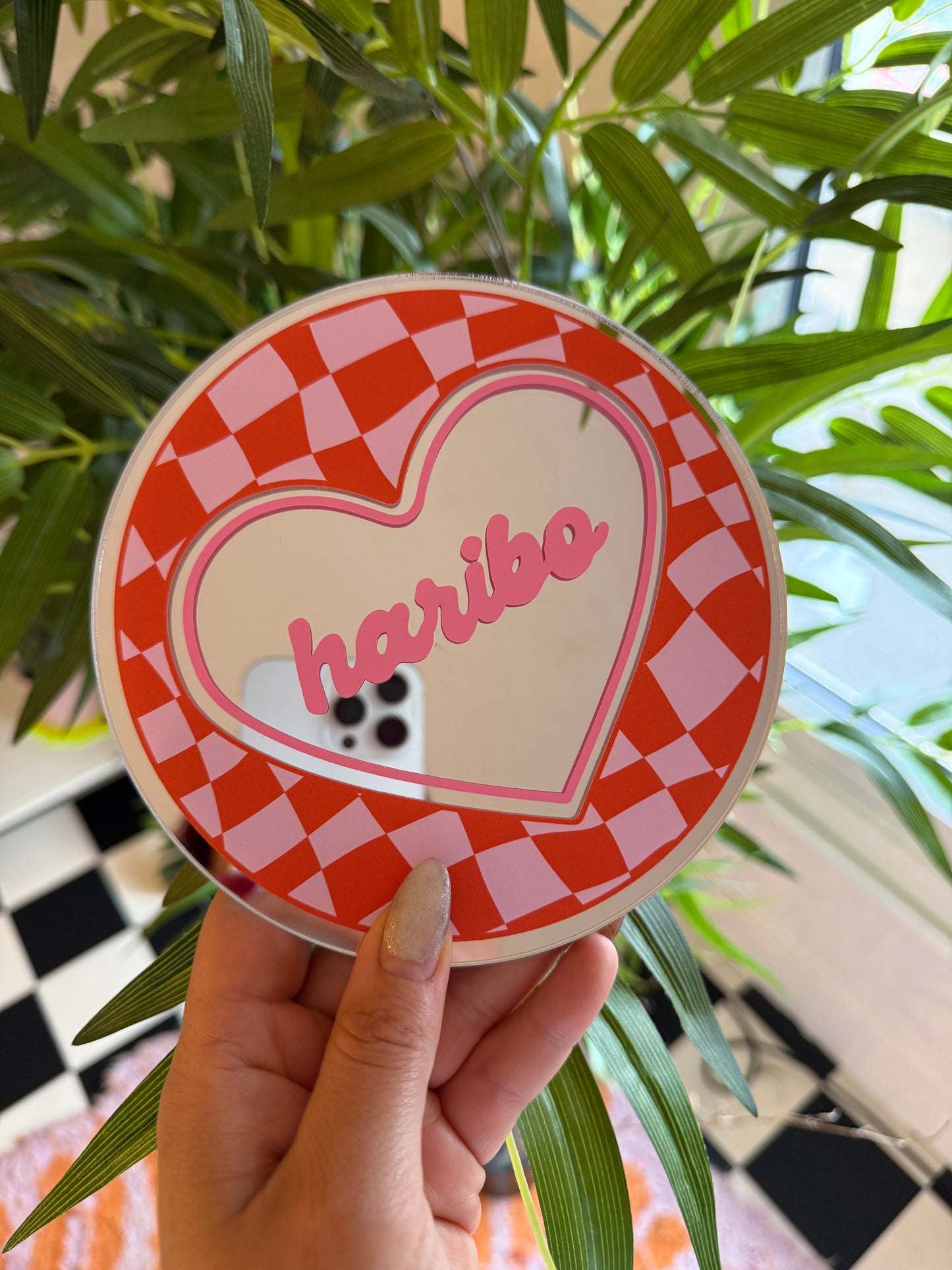 A hand is holding a heart-shaped object with the word "Love" written on it, against a backdrop of green leaves and a checkered pattern.
