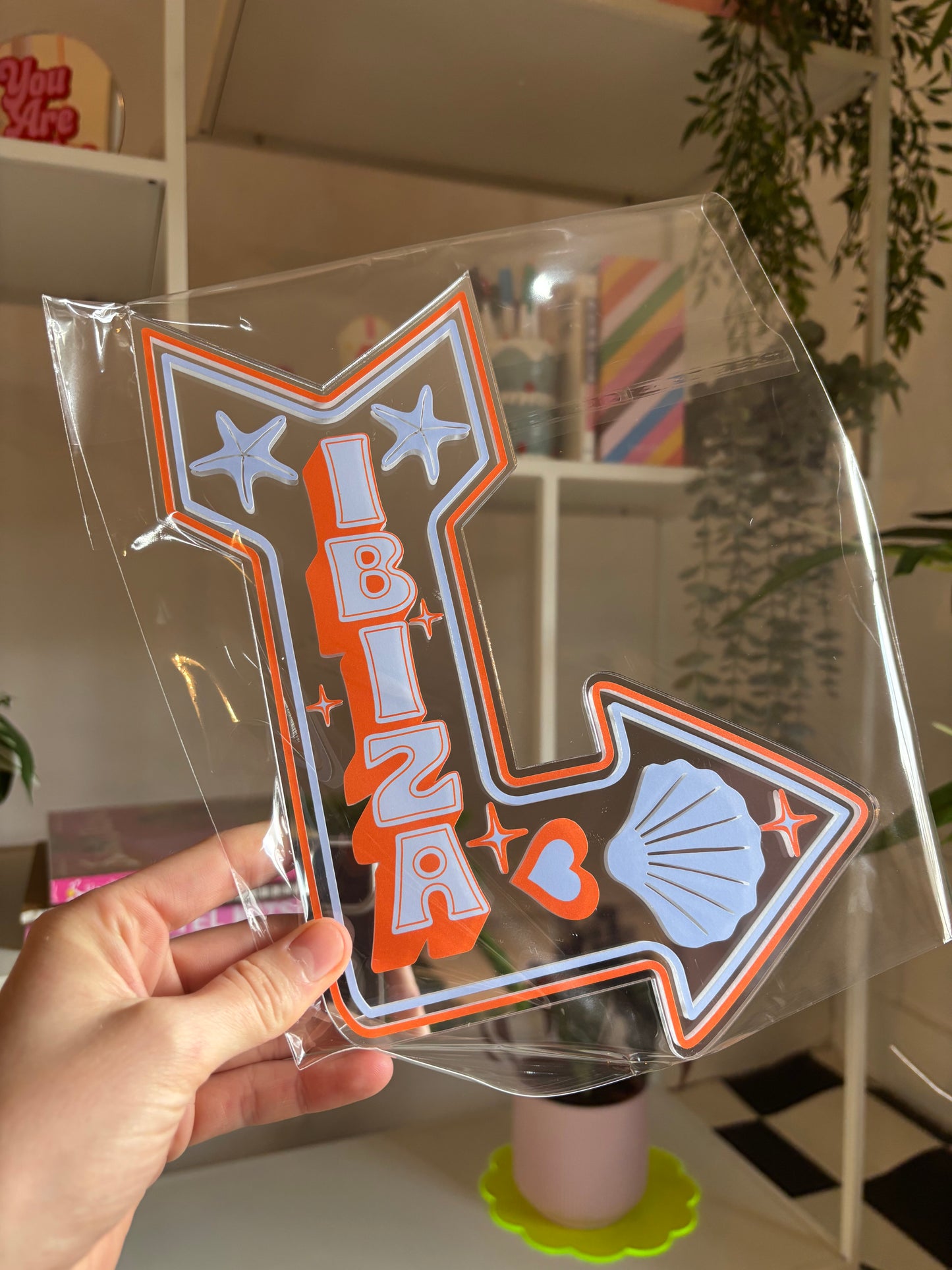 A hand is holding a clear plastic sign with the word "IBIZA" written on it, along with various decorative elements such as stars, hearts, and a shell.