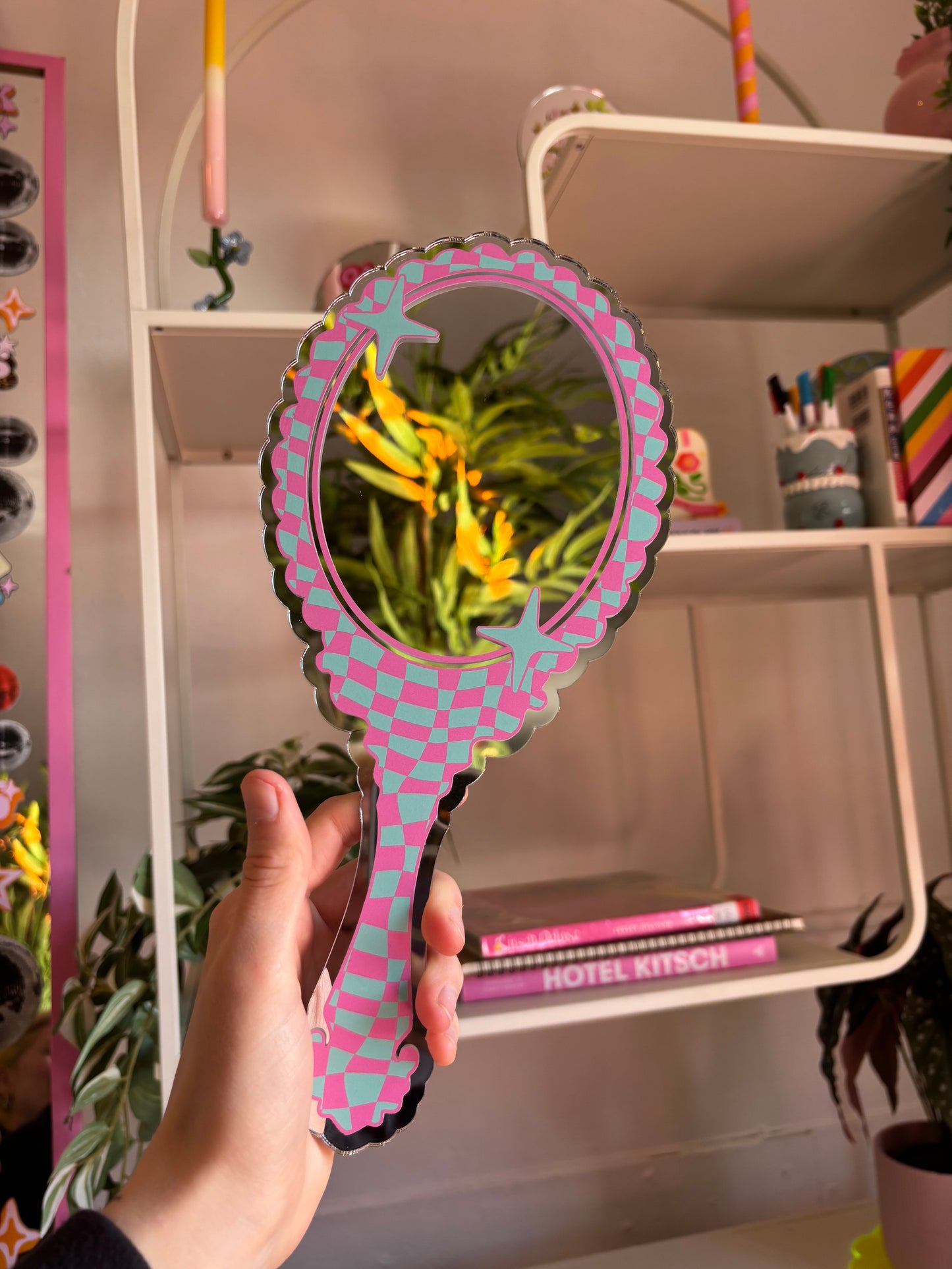 A hand holding a pink and green checkered mirror with a plant in the background.