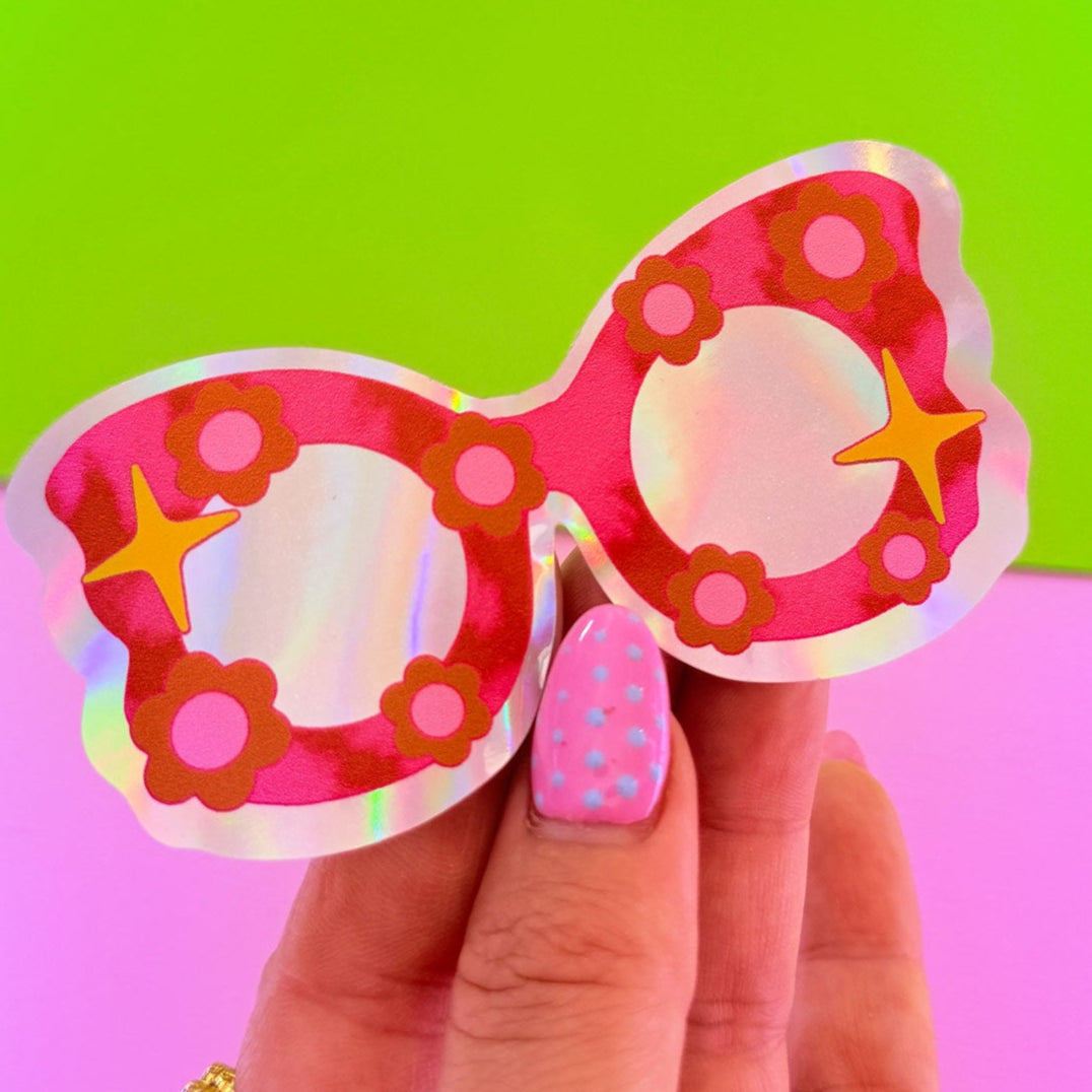 Hand holding a colorful sunglasses sticker against a green and pink gradient background