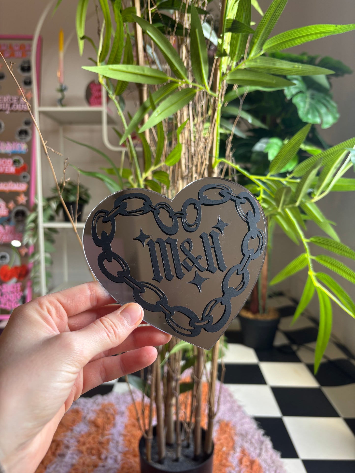 A hand is holding a heart-shaped sticker with the text "M&H" on it, in front of a potted plant with green leaves.
