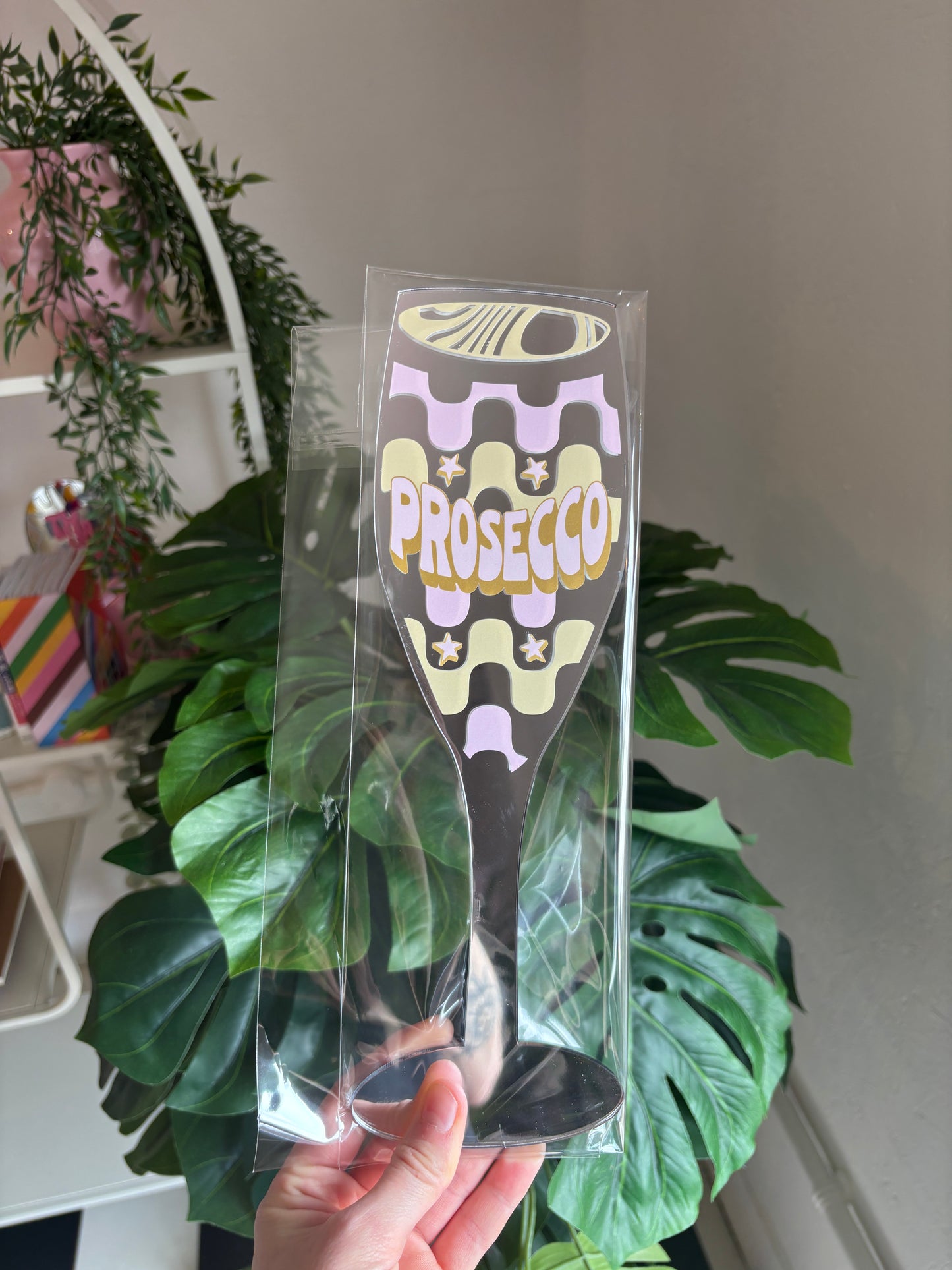 A hand is holding a clear plastic object with a decorative design, which appears to be a glass or a container with a label or text on it. The background features a large green plant.