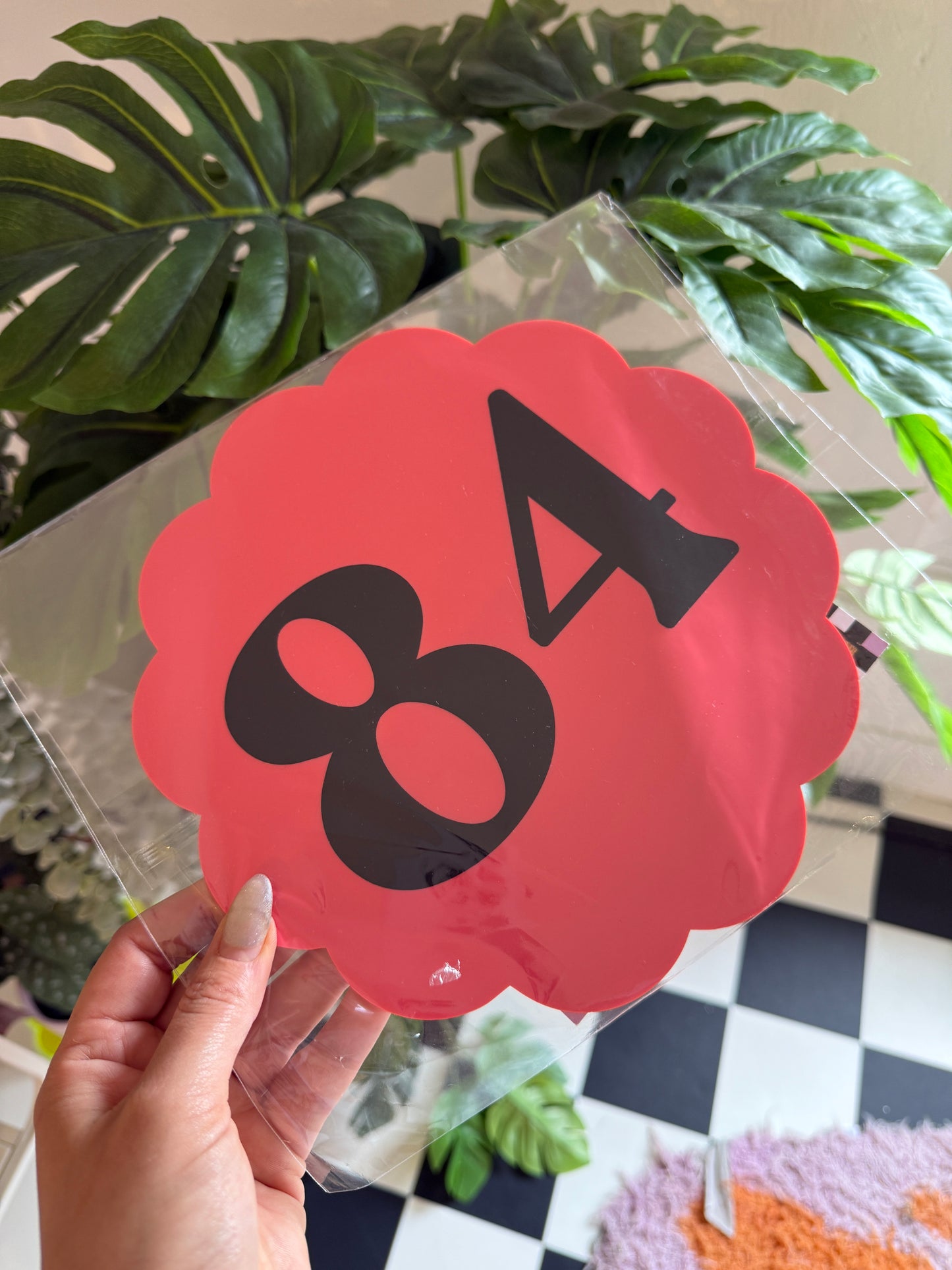 A hand is holding a clear plastic sheet with a red and black number 8 on it, against a backdrop of green plants and a checkered wall.