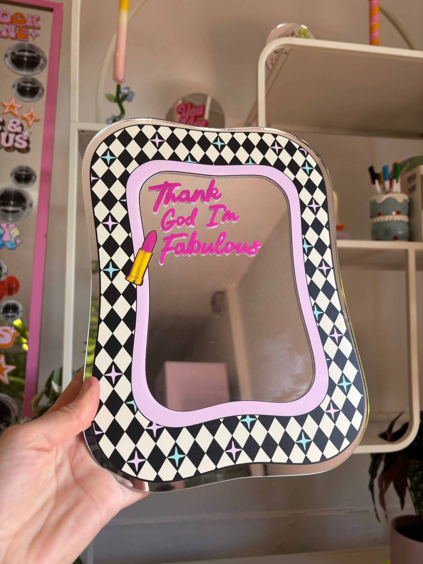 A hand holding a mirror with a black and white checkered frame and a pink and purple border. The mirror has a message written on it that says "Thank God I'm Fabulous".