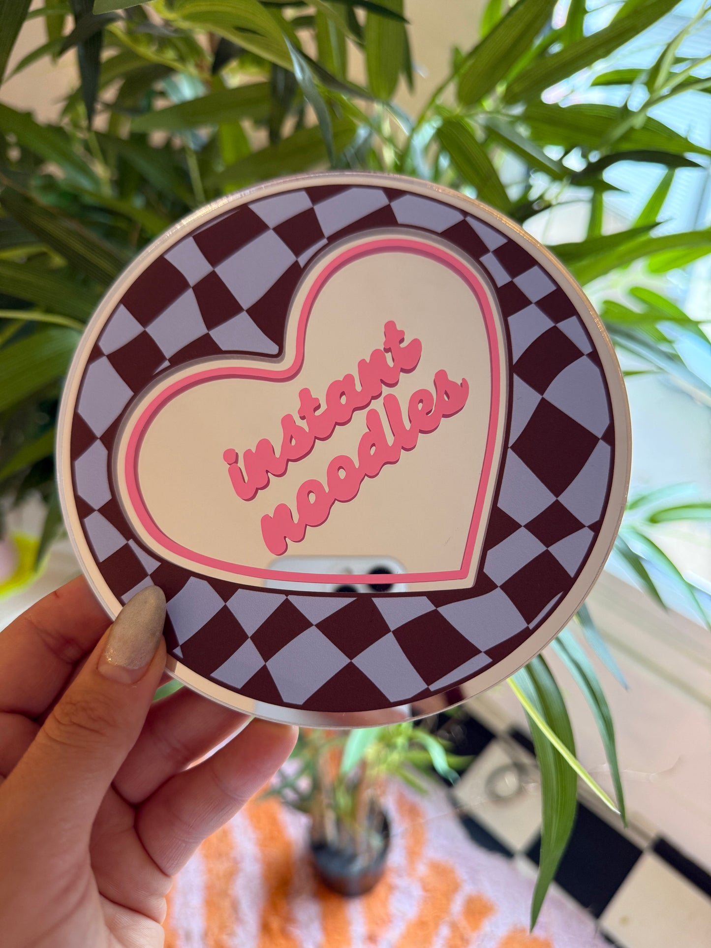 A hand holding a heart-shaped object with a checkered pattern and pink text on it, in front of a plant.