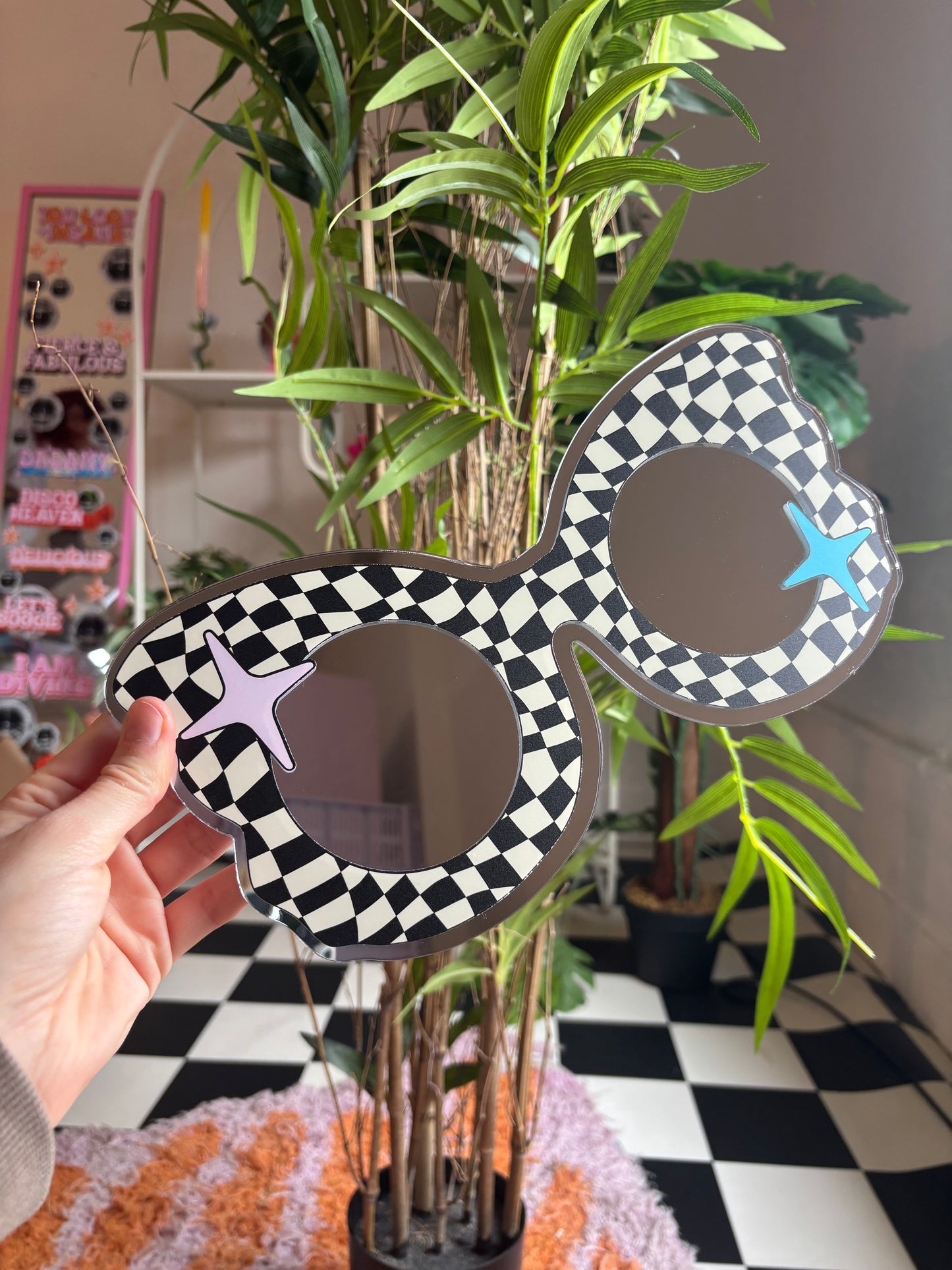 A hand is holding a pair of black and white checkered sunglasses with blue stars, which are placed in front of a potted plant.