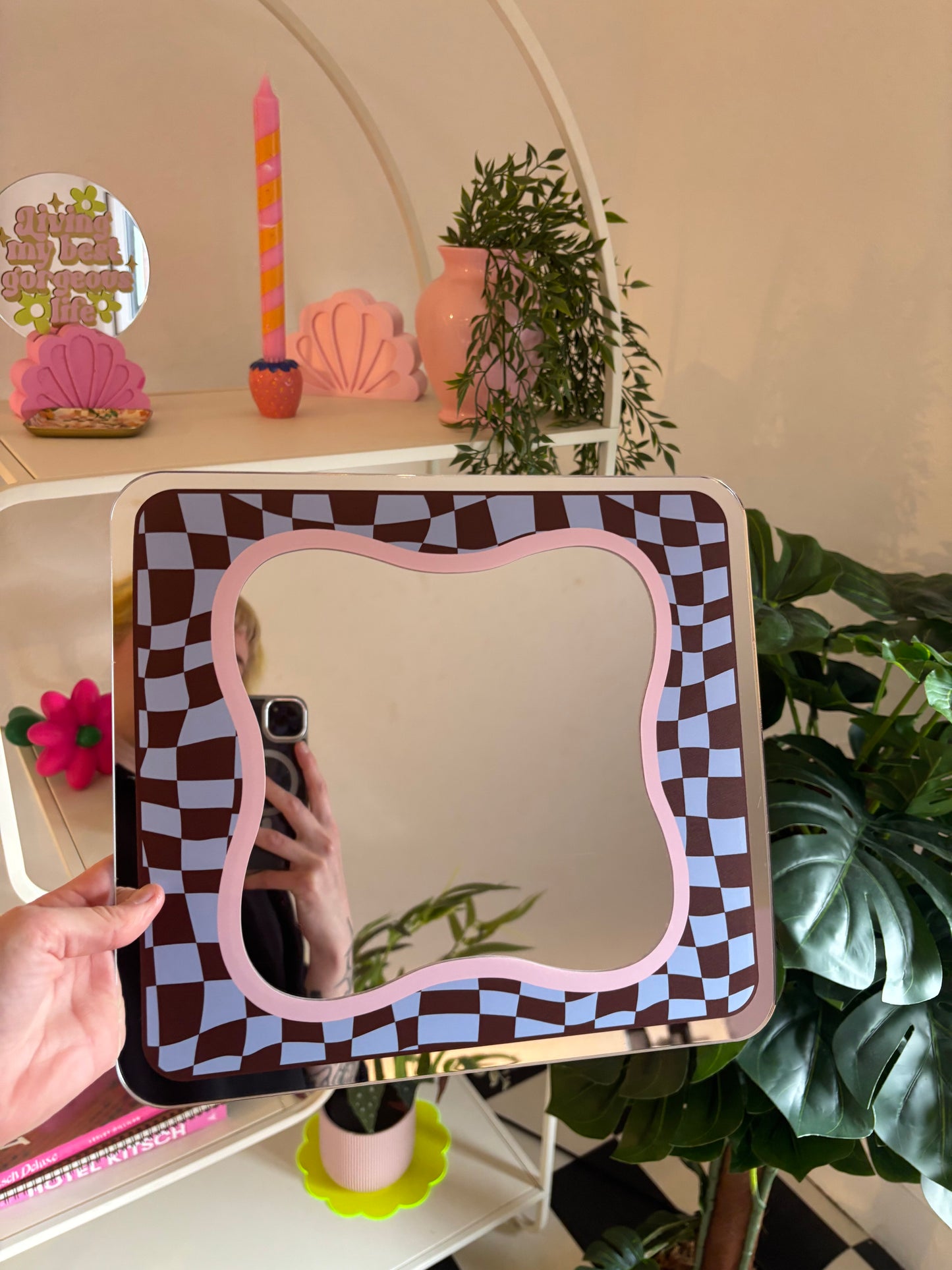 A hand is holding a mirror with a checkered frame, which is placed on a shelf with various decorative items such as a candle, a vase, and a potted plant.