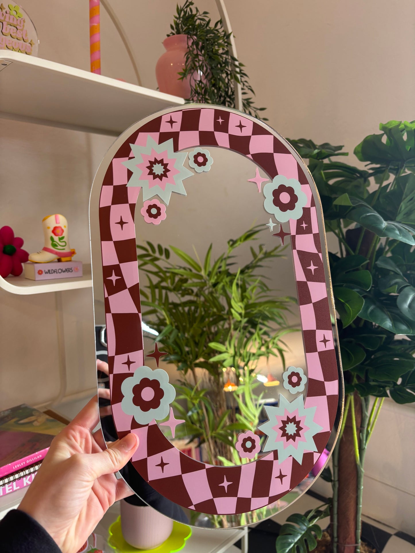 A hand is holding a mirror with a checkered frame and floral designs, with a shelf of decorative items and plants in the background.