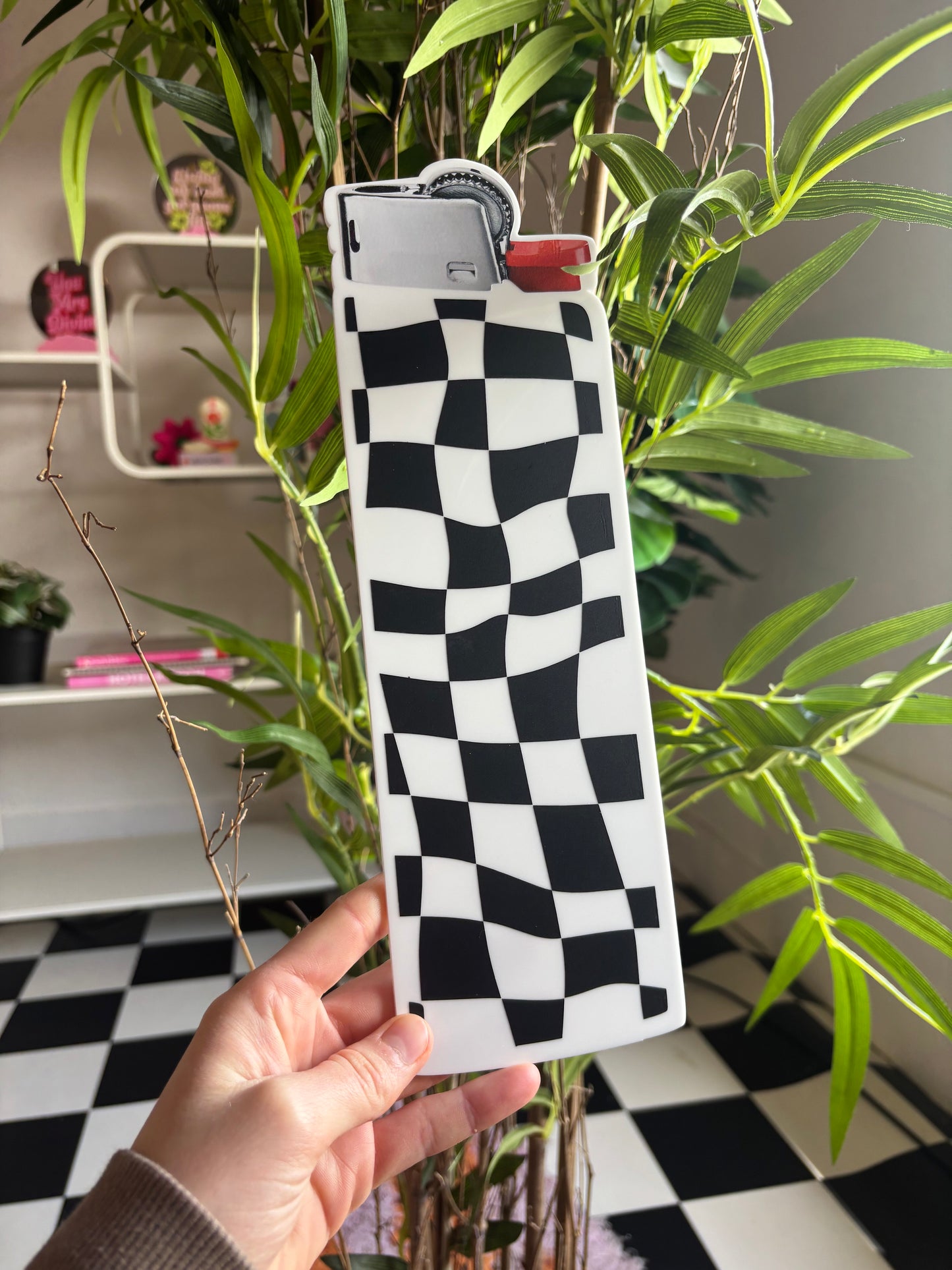 A hand is holding a black and white checkered lighter with a red lighter attached to it, standing in front of a plant with green leaves.
