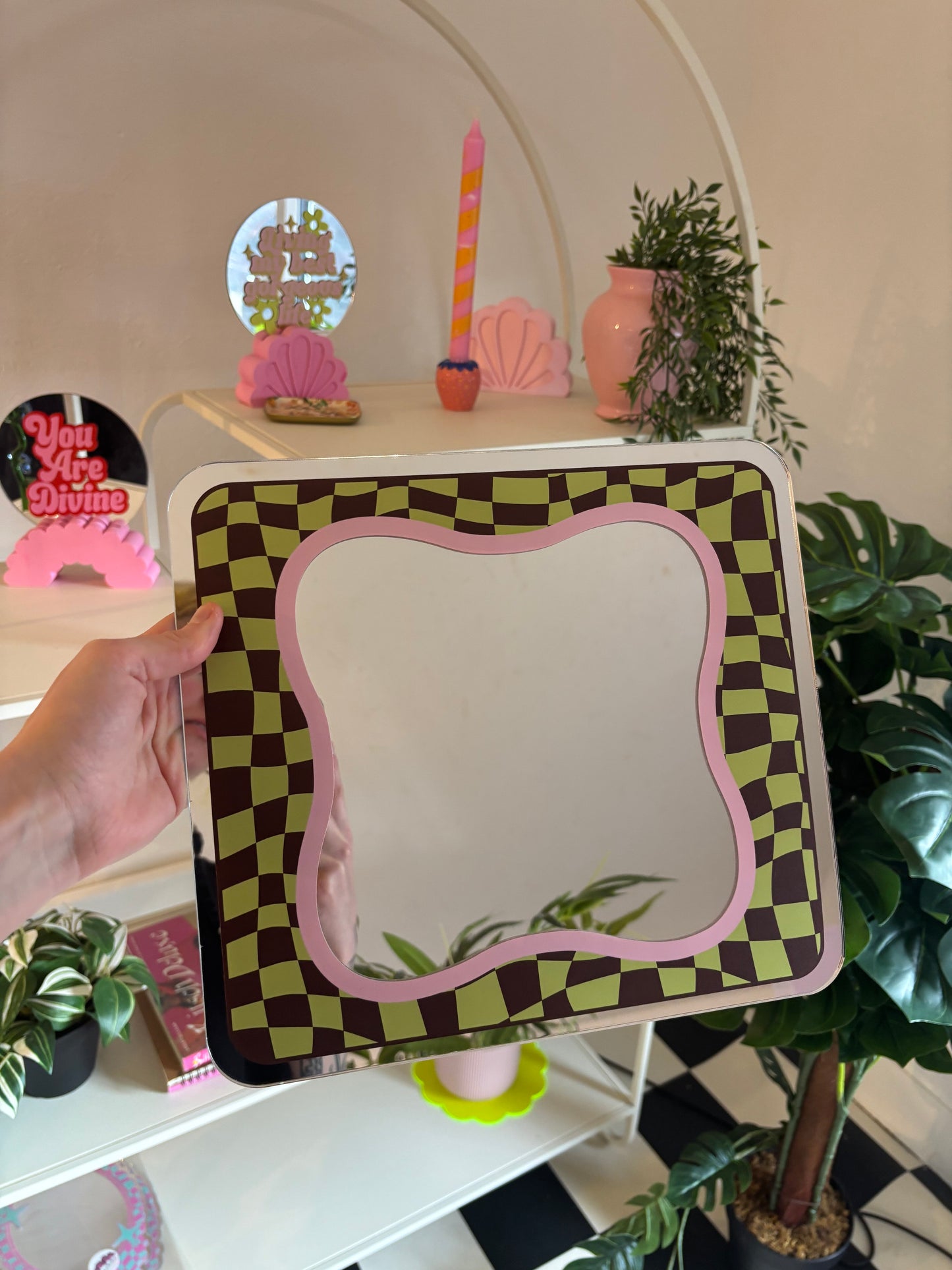 A hand is holding a mirror with a checkered frame, which is placed on a shelf among various decorative items.