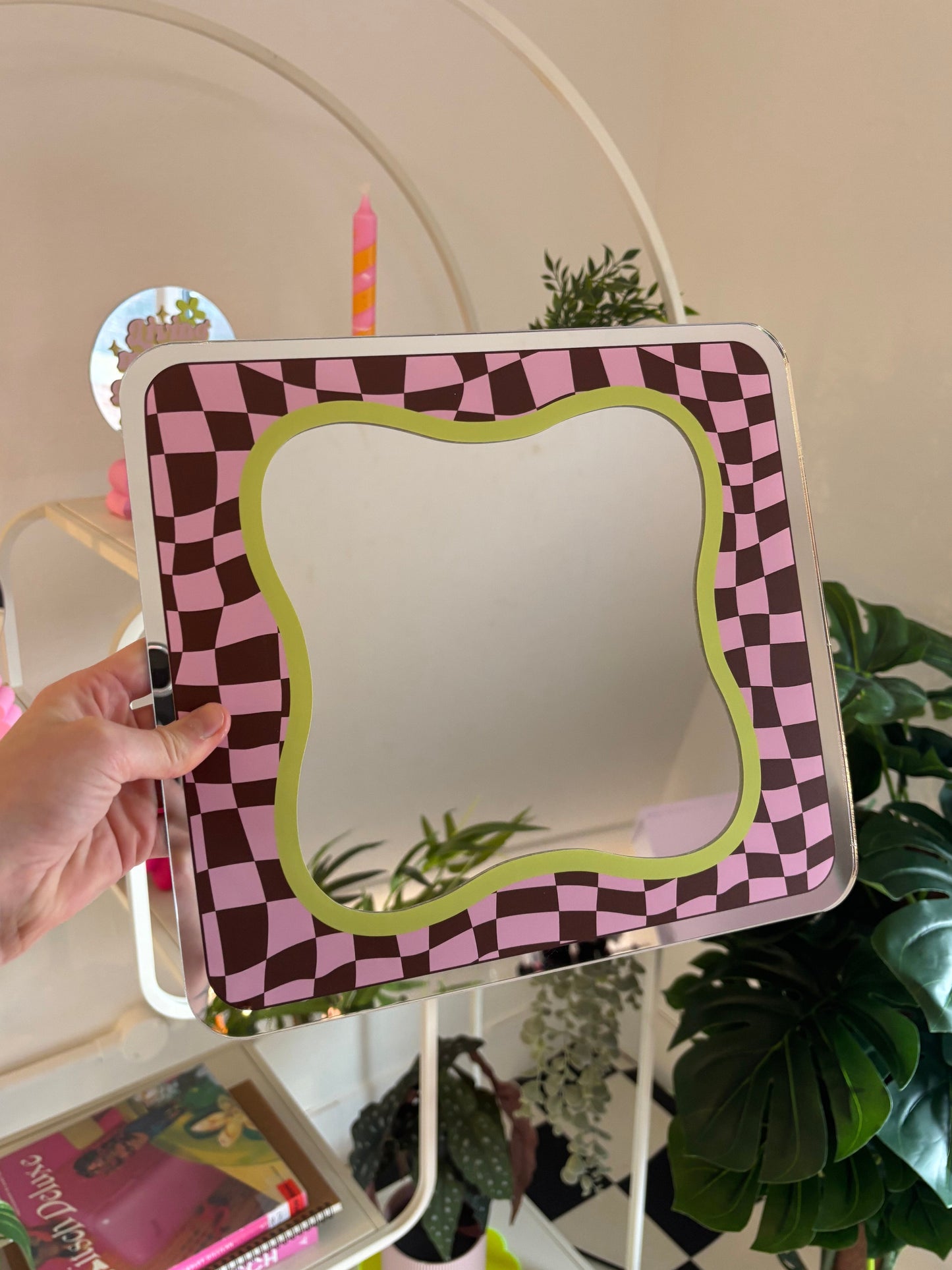 A hand is holding a square mirror with a checkered frame in a room with various plants and decor.