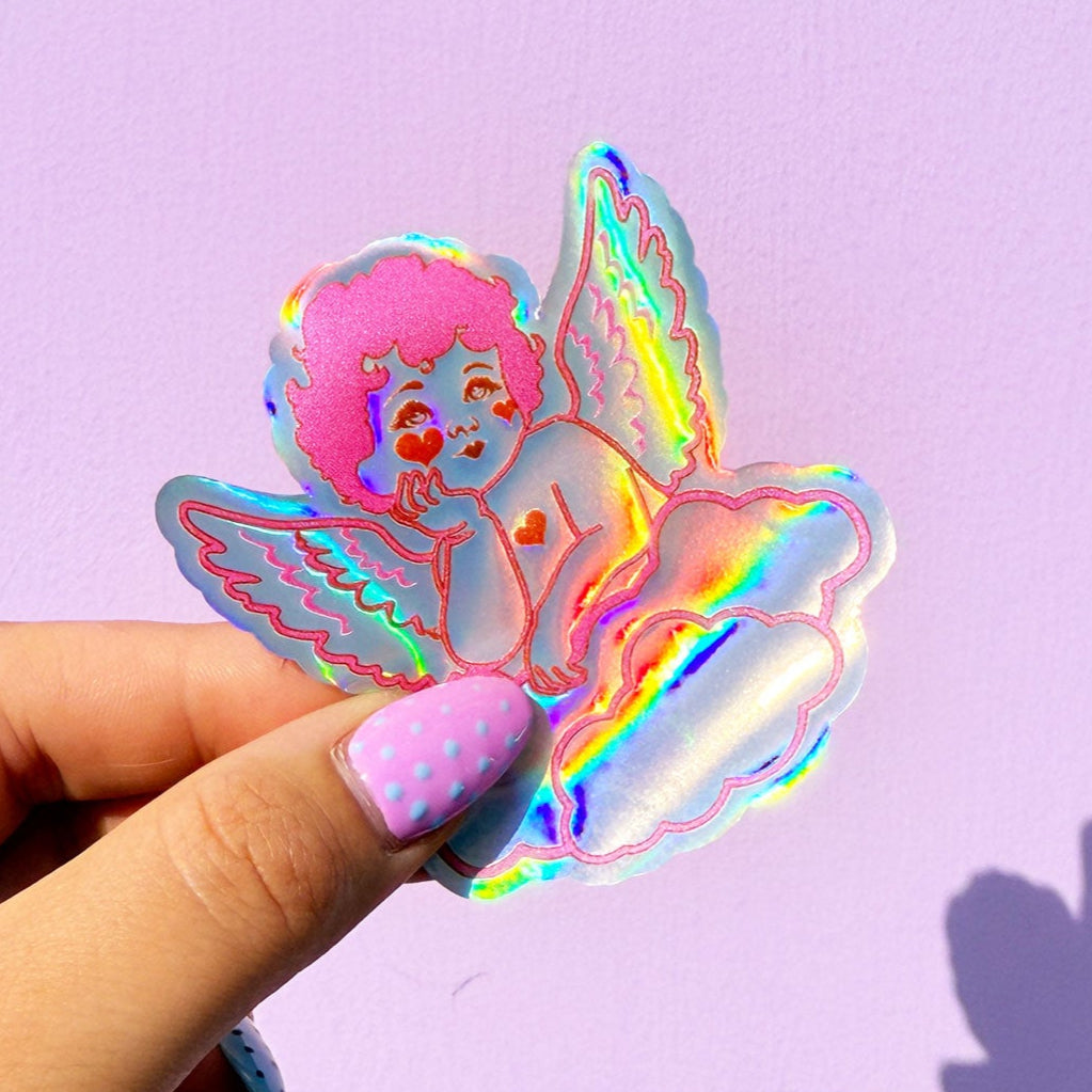 Holographic angel sticker held by a hand with pink nail polish against a purple background