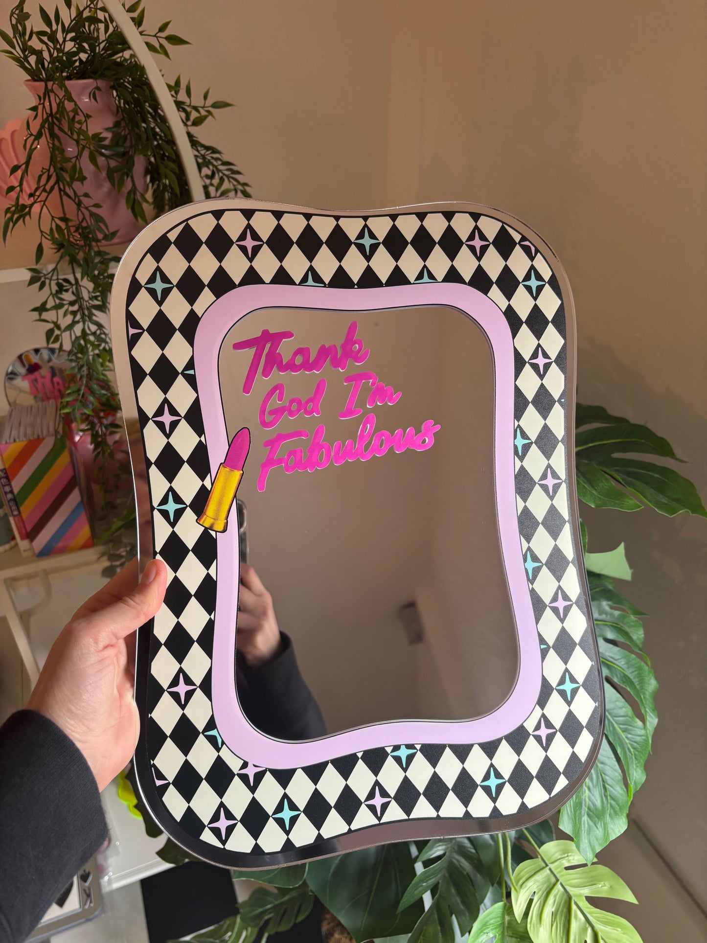 A hand holding a mirror with a decorative frame, featuring a pink lipstick and a checkered pattern.