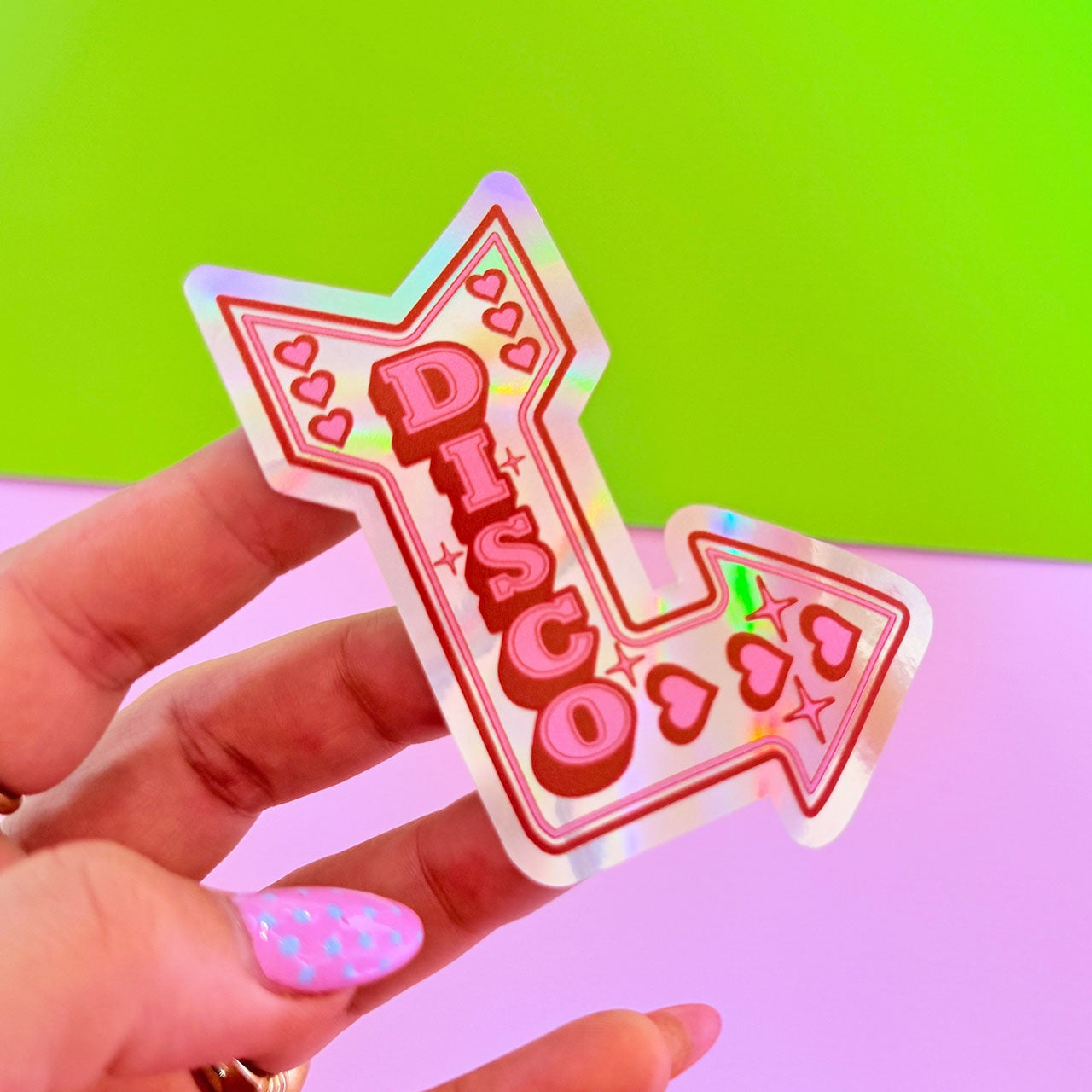 Holographic 'DISCO' sticker held by a hand with pink nail polish against a green and pink gradient background.