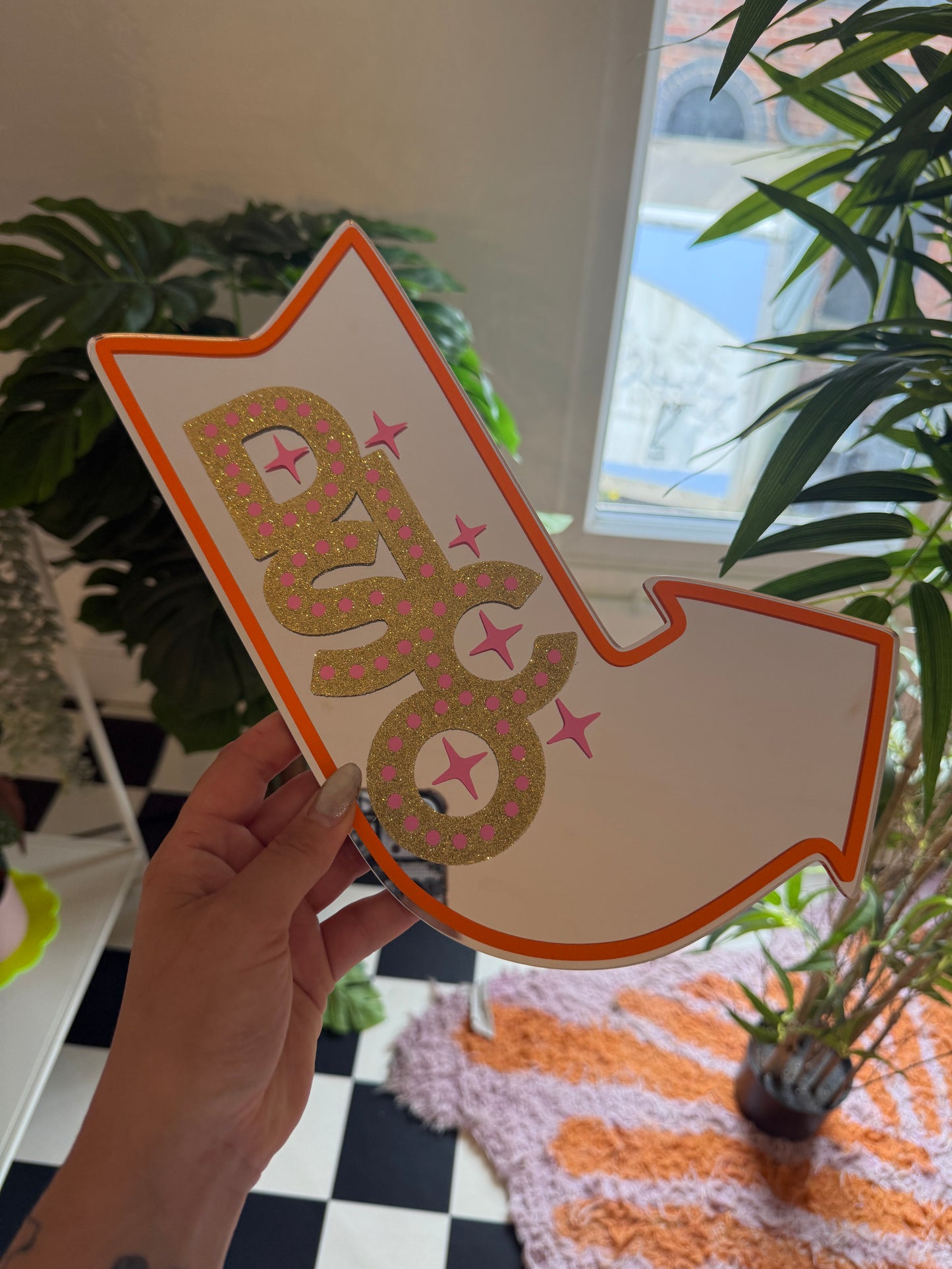 A hand is holding a white arrow-shaped object with pink and gold glitter and stars on it, which appears to be a decorative piece.
