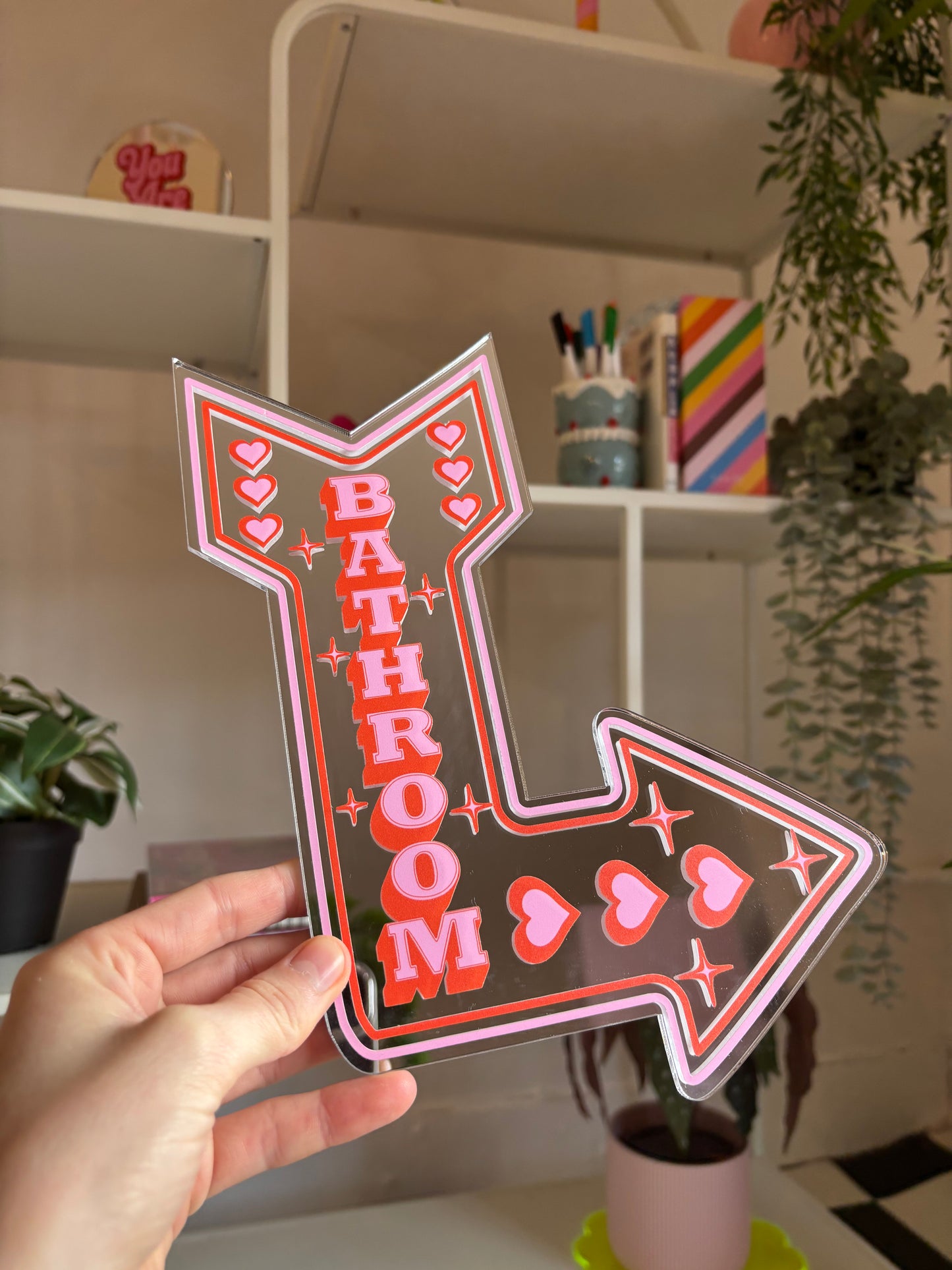 A hand is holding a large, colorful sign that says "BATHROOM" in bold letters, with hearts and stars decorating the sign.