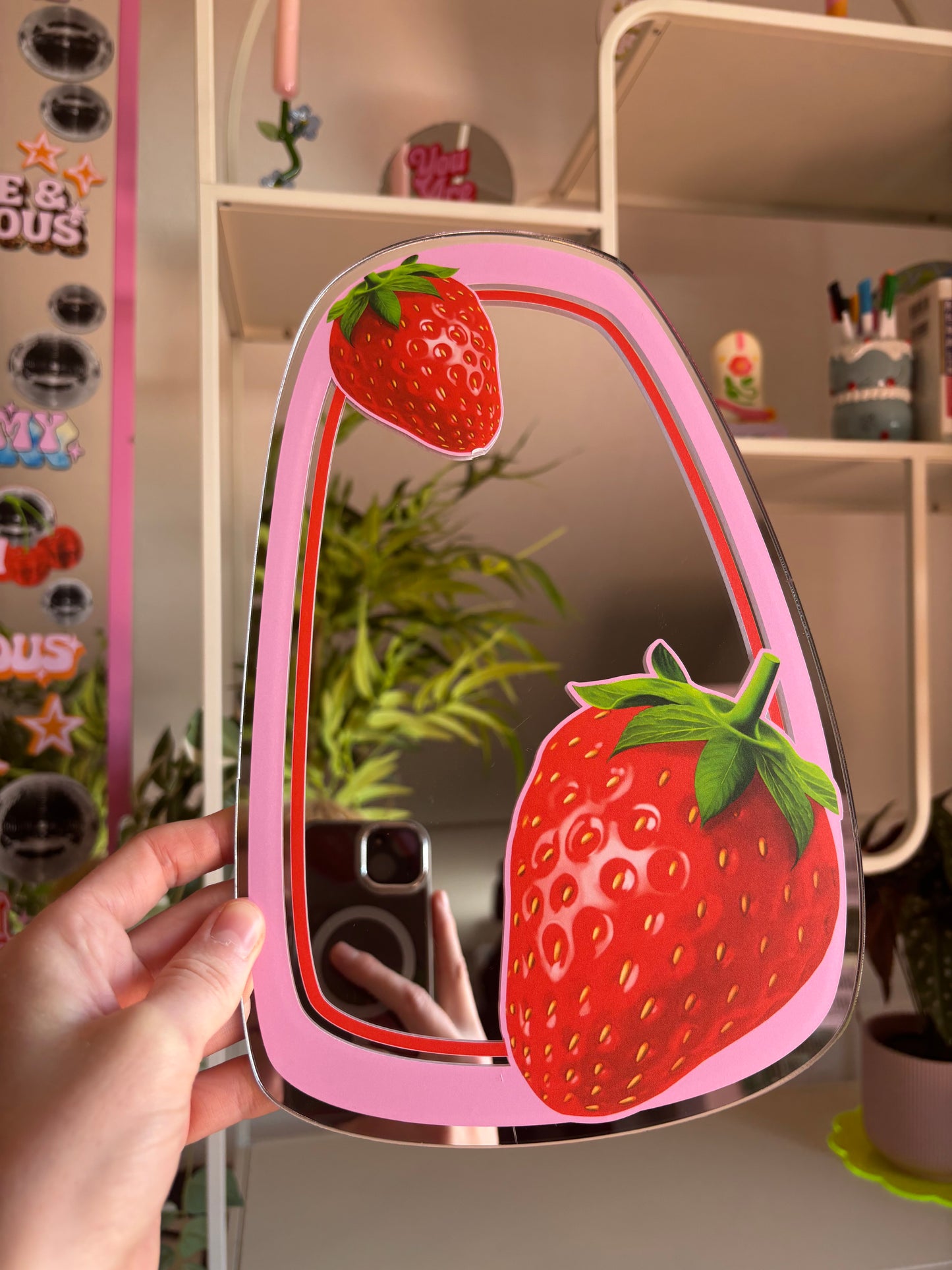 A hand is holding a pink mirror with a strawberry design, and the reflection of a person taking a photo of the mirror can be seen in the background.