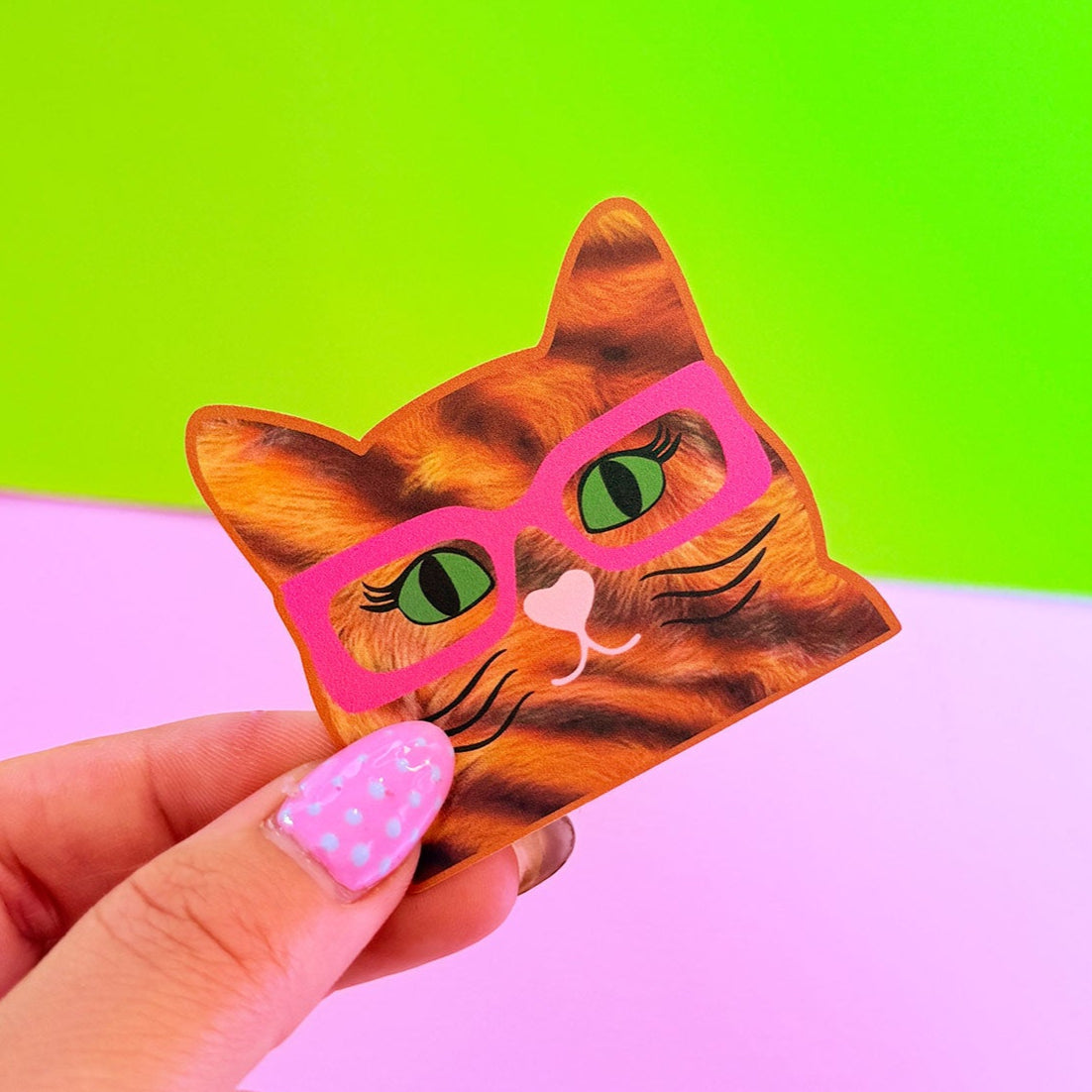 Hand holding a cat-shaped card with pink glasses against a bright green and pink background