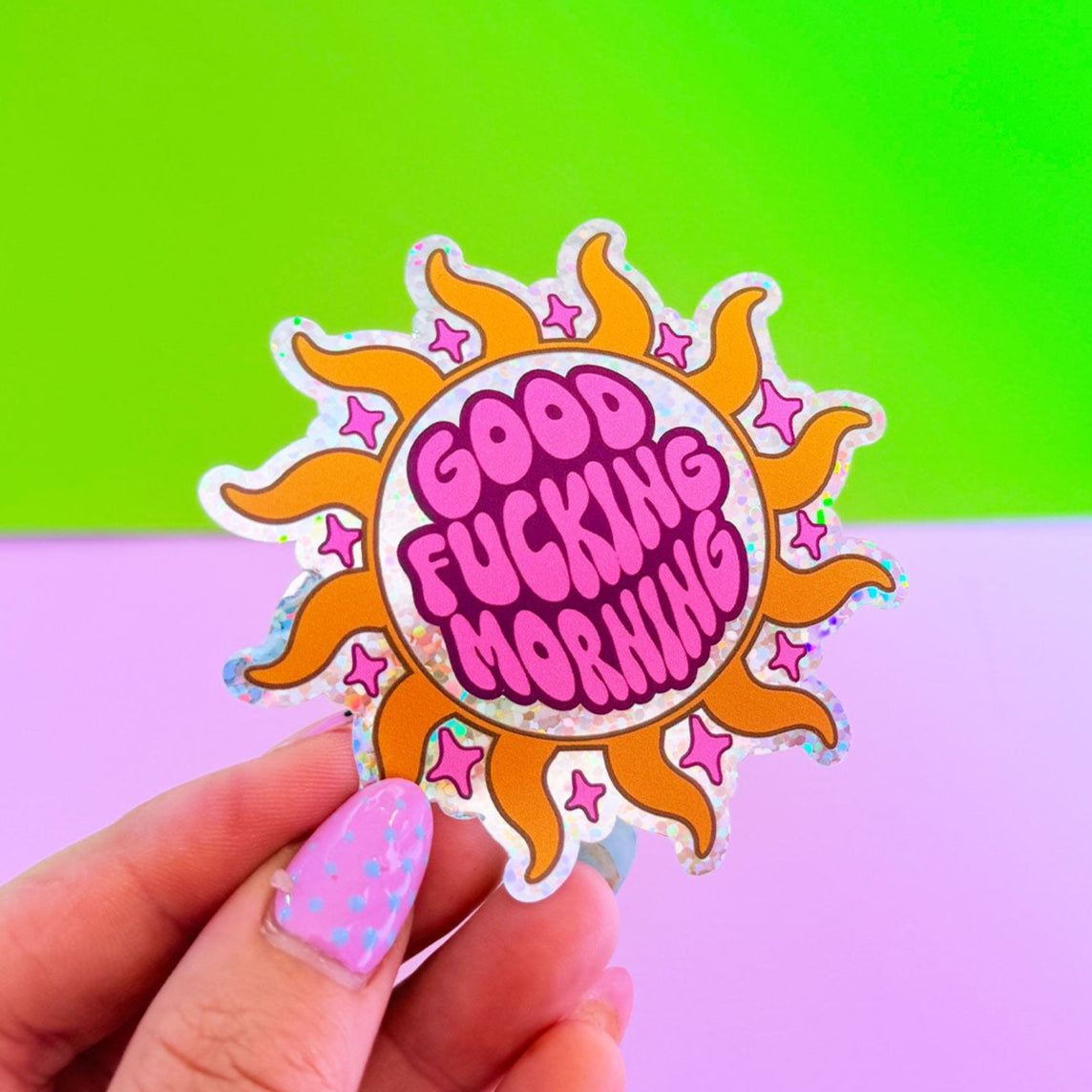 Hand holding a colorful sticker with 'Good Fucking Morning' text against a green and pink gradient background.