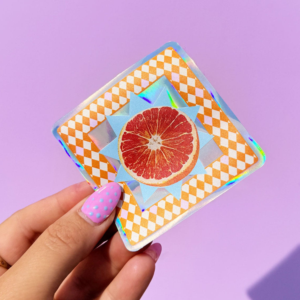 Hand holding a square sticker with a grapefruit design on a purple background