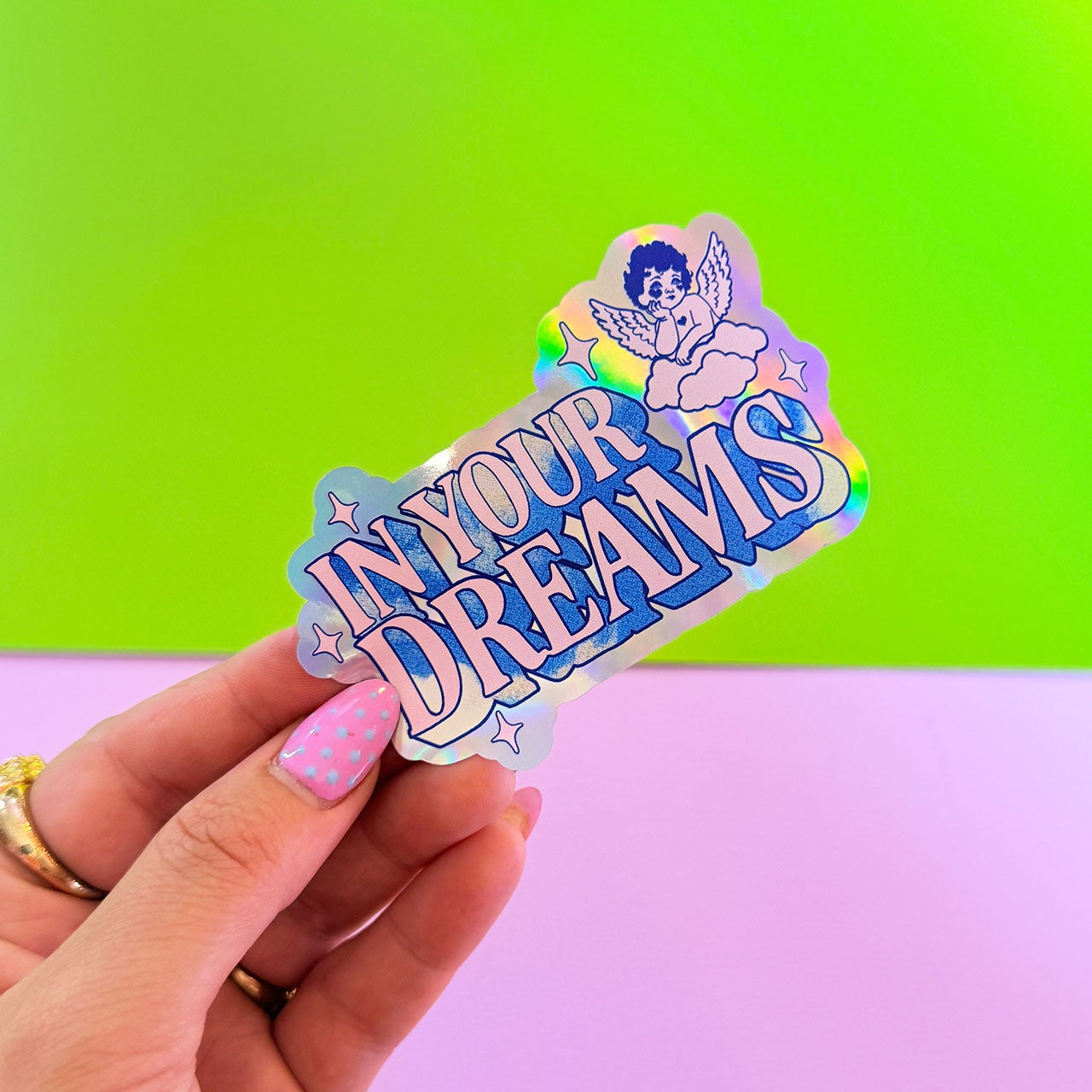 Hand holding a colorful sticker with 'In Your Dreams' text against a green and pink gradient background