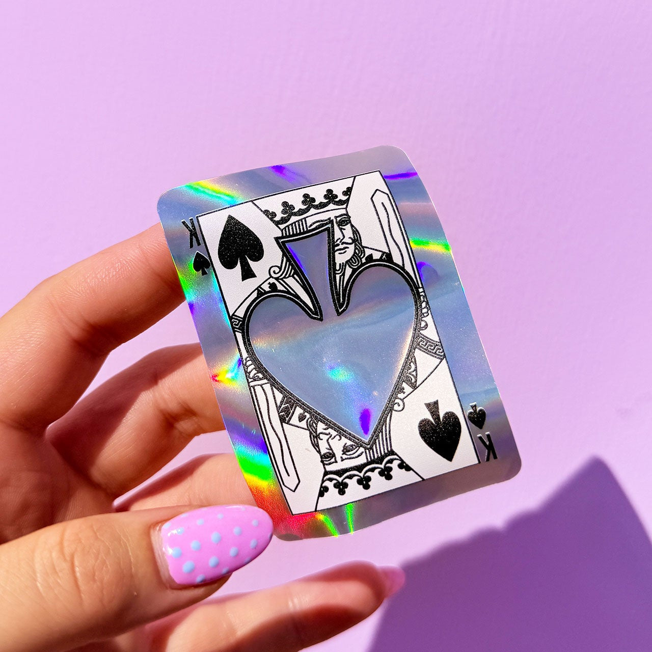 Holographic King of Hearts playing card held by a hand with pink nail polish against a purple background