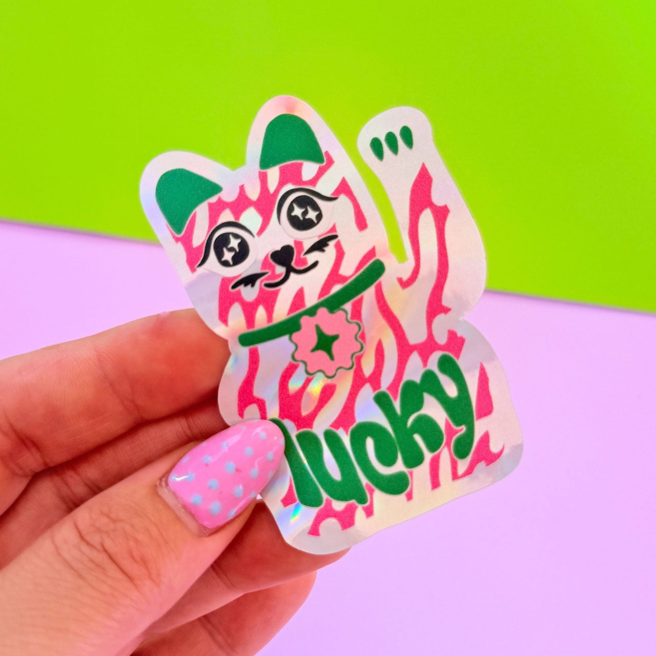 Sticker of a pink cat with green accents and 'lucky' text held by a hand against a colorful background