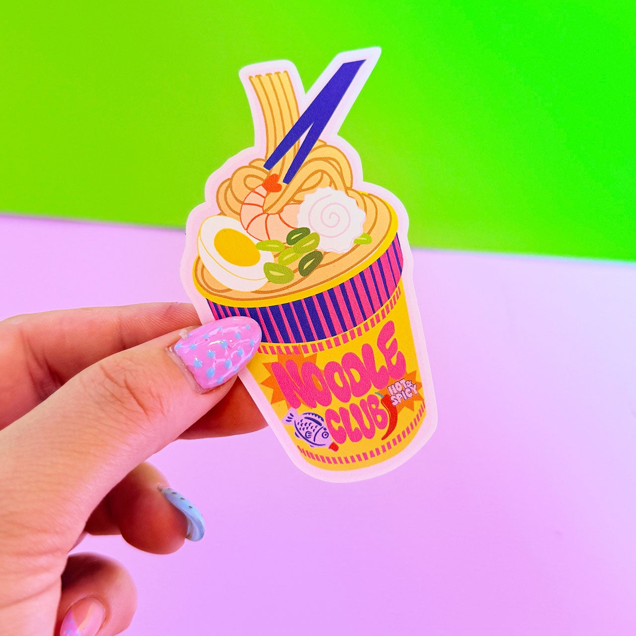 Hand holding a colorful noodle cup toy against a green and pink gradient background