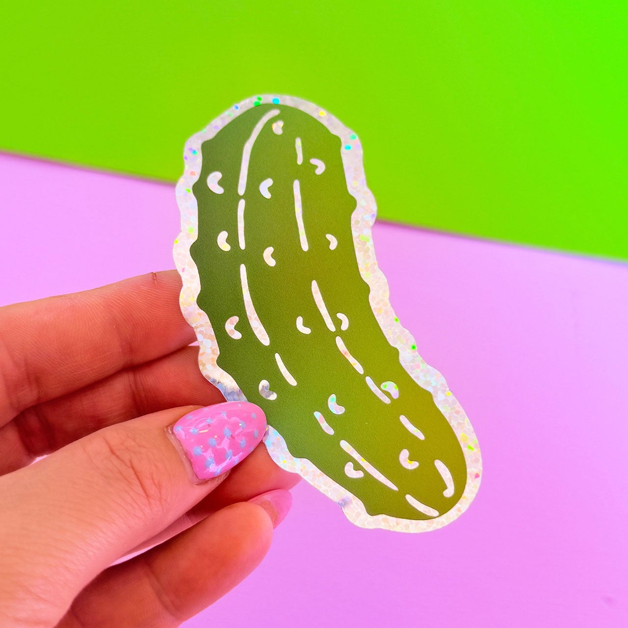 Hand holding a pickle-shaped sticker against a green and pink background