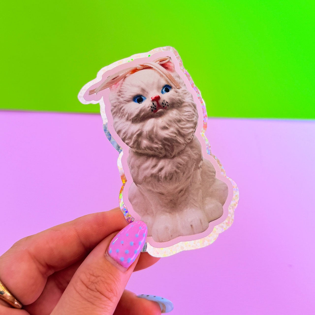 Sticker of a cat with bunny ears held by a hand against a colorful background