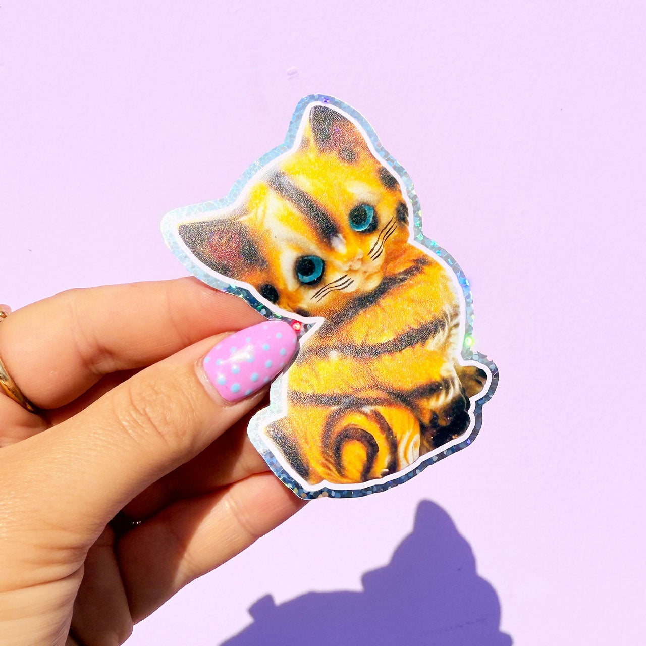 Holographic cat sticker held by a hand with pink nail polish on a purple background