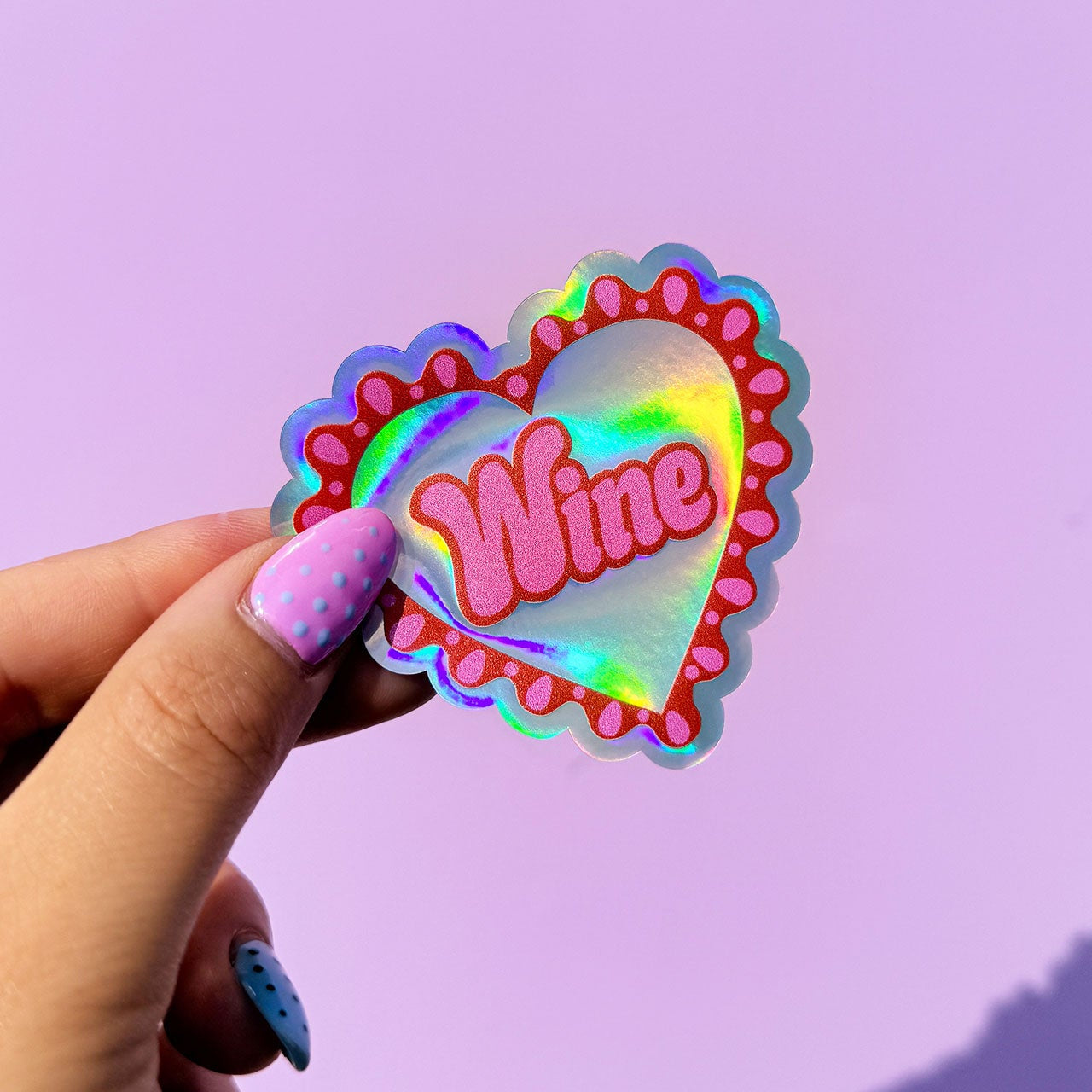 Holographic 'Wine' heart-shaped sticker held by a hand against a purple background